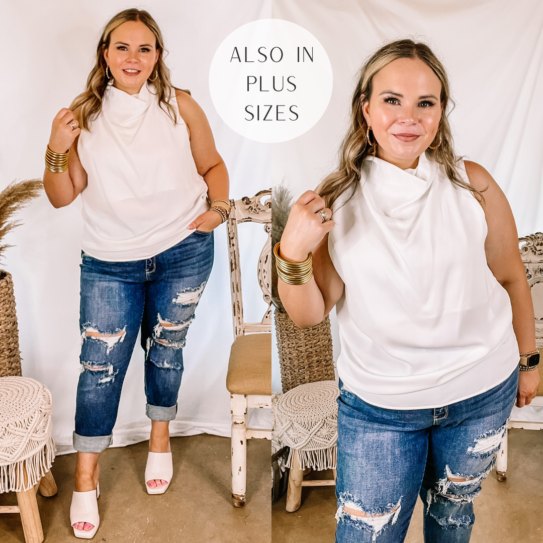 Model is wearing a white cowl neck tank top. Model has it paired with distressed boyfriend jeans, white heels, and gold jewelry.