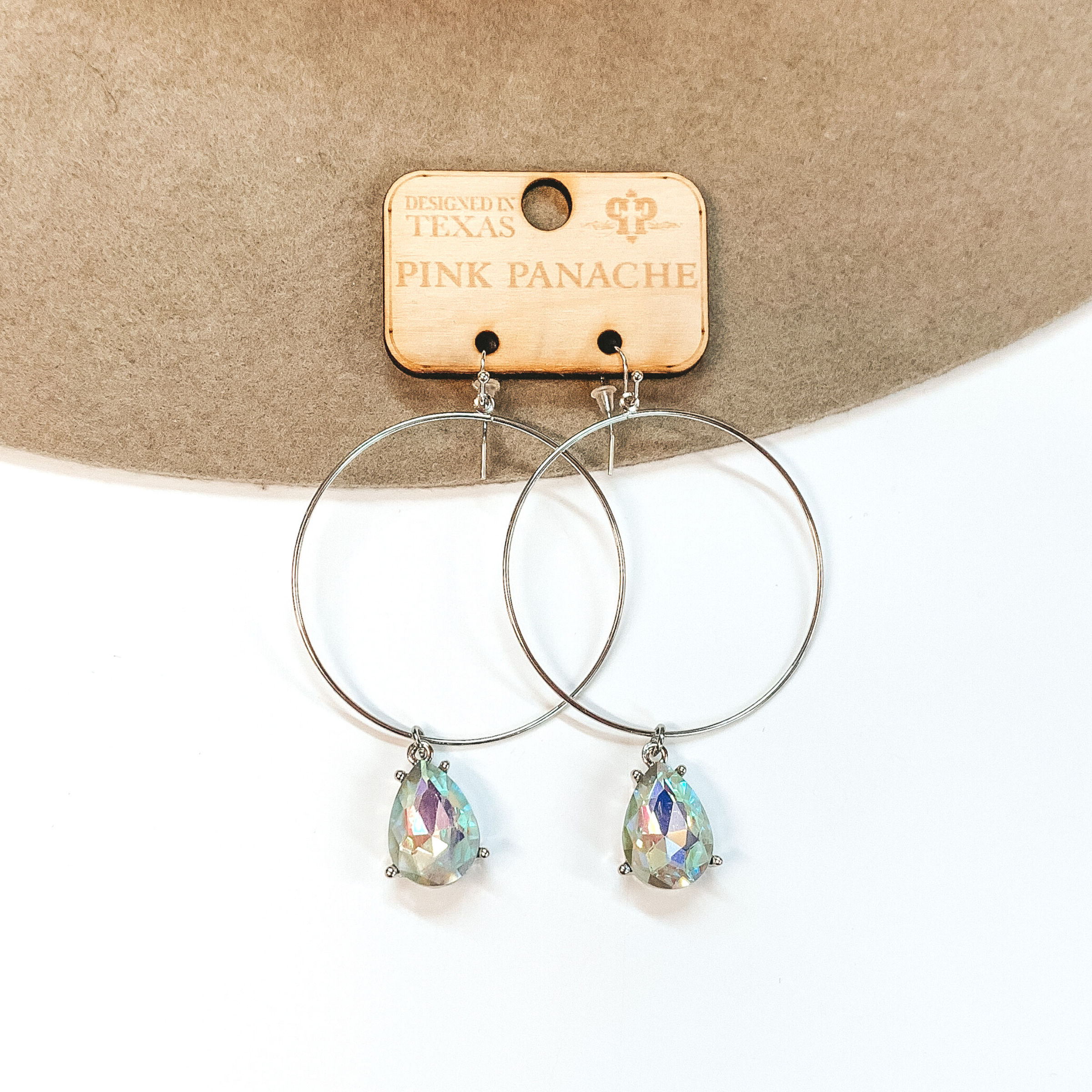 Silver hoop earrings with a teardrop ab crystal dangle. Pictured with a beige hat on a white background.