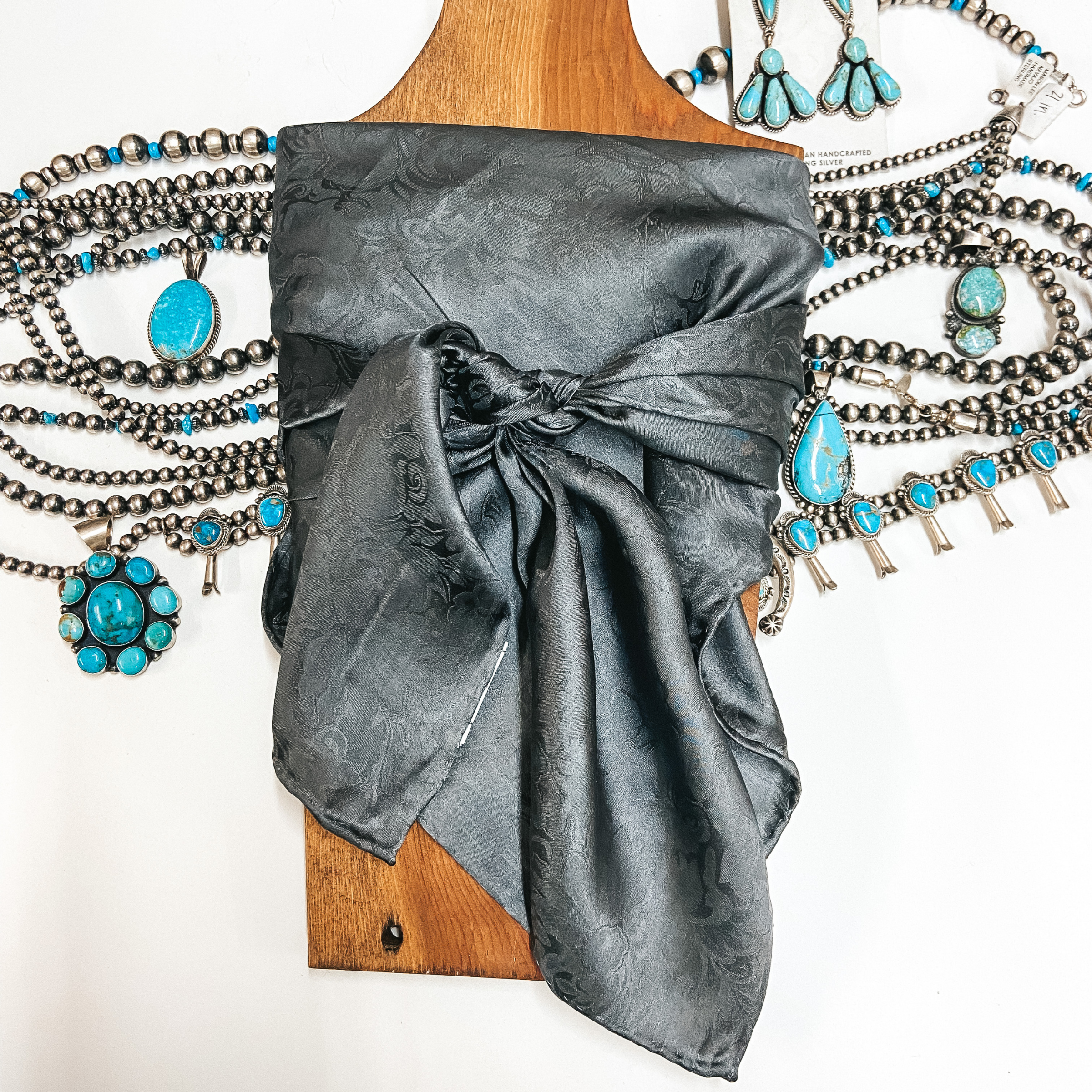 A jacquard print wild rag tied around a wooden display. Pictured on white background with sterling silver and turquoise jewelry.
