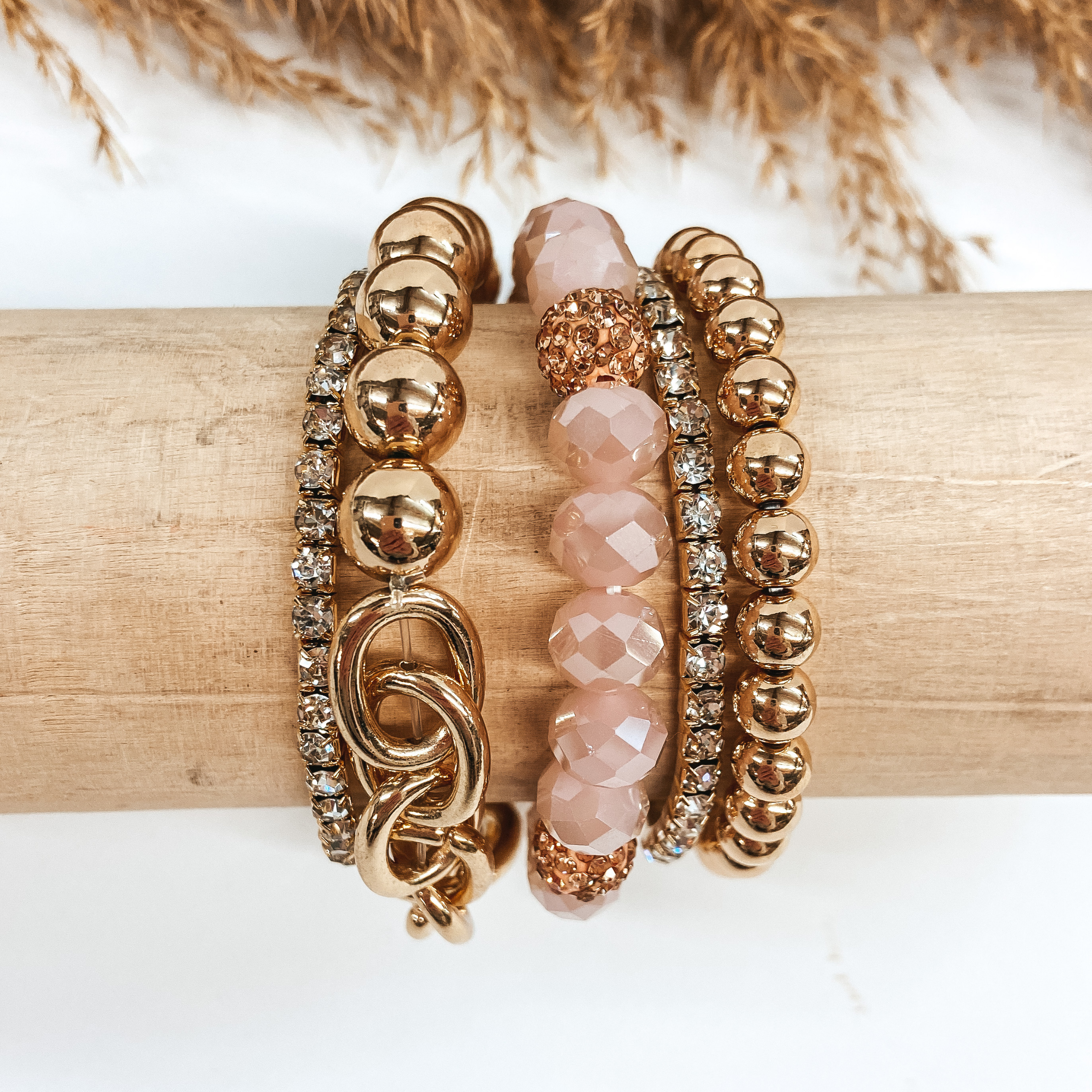 A mix of crystal and gold beaded bracelets placed on a wooden slat. Pictured on a white background with pampas grass.
