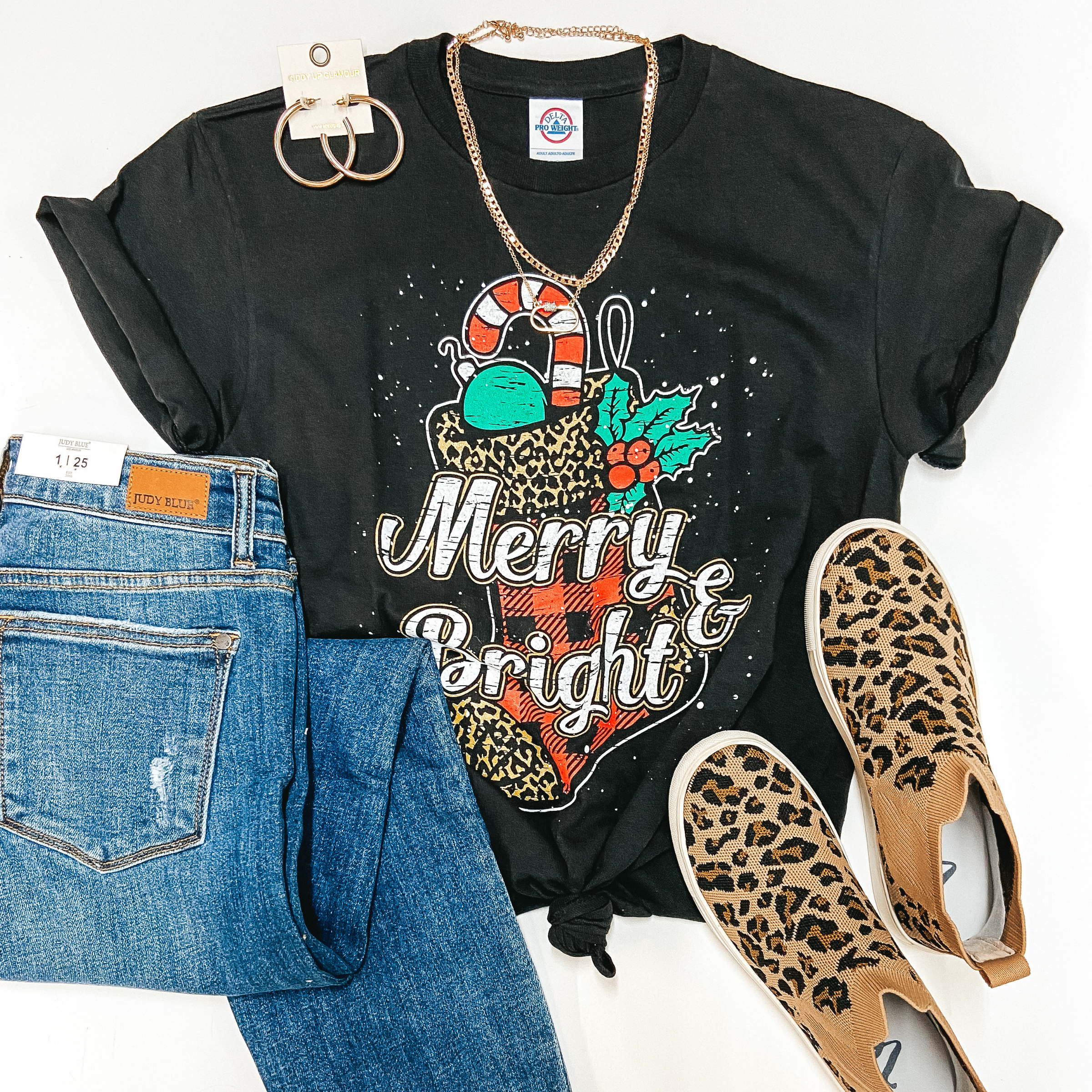 A black tee shirt with cuffed sleeves and knotted front. The tee has a graphic of a leopard stocking and says "Merry and Bright." Pictured on a white background with medium wash jeans, high top leopard sneakers, and gold jewelry.