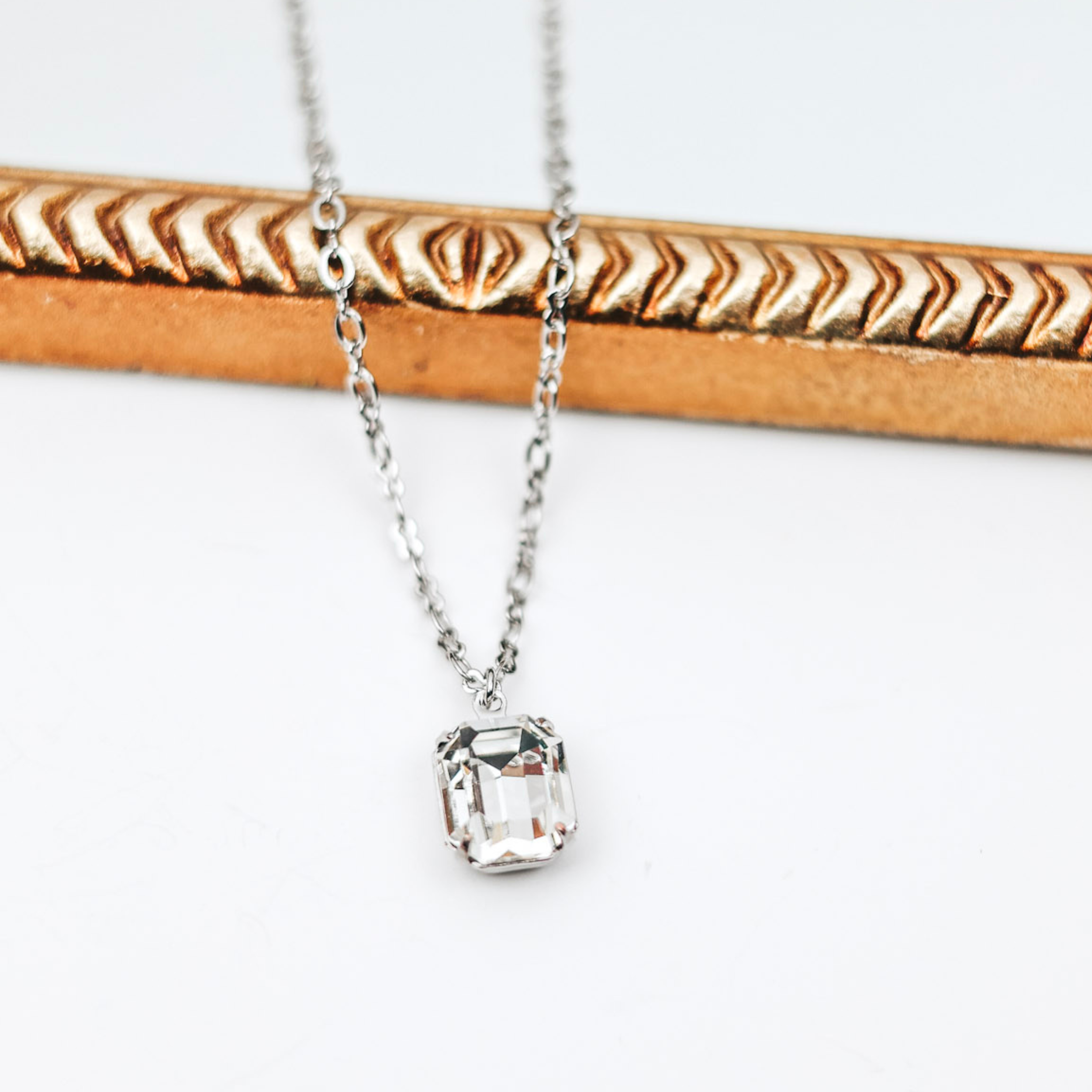 A silver-tone necklace with a single crystal pendant that is emerald cut. This necklace is pictured on a white background with a mirror.