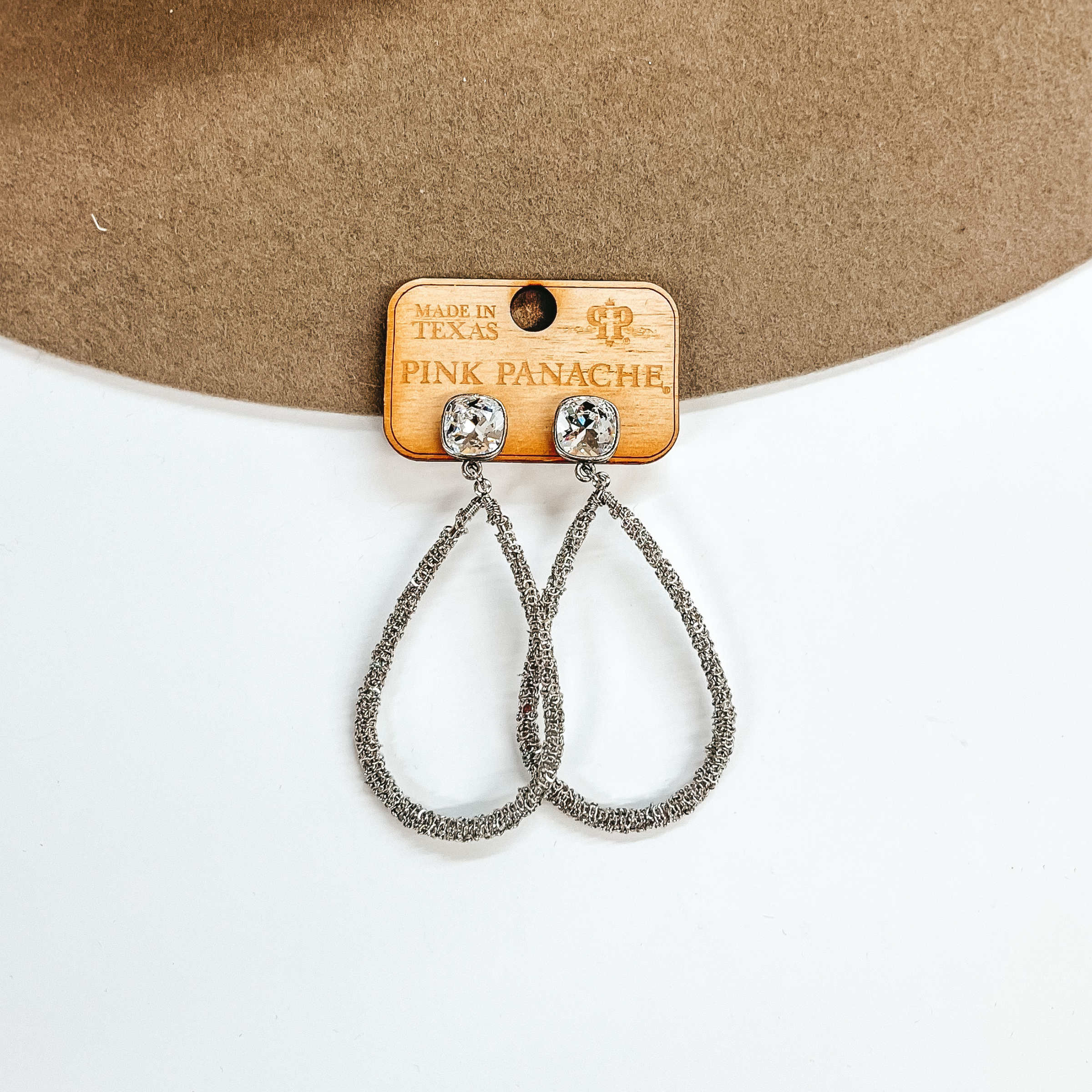 Silver teardrop earrings with a clear cushion cut crystal. Pictured on white background with a beige hat.