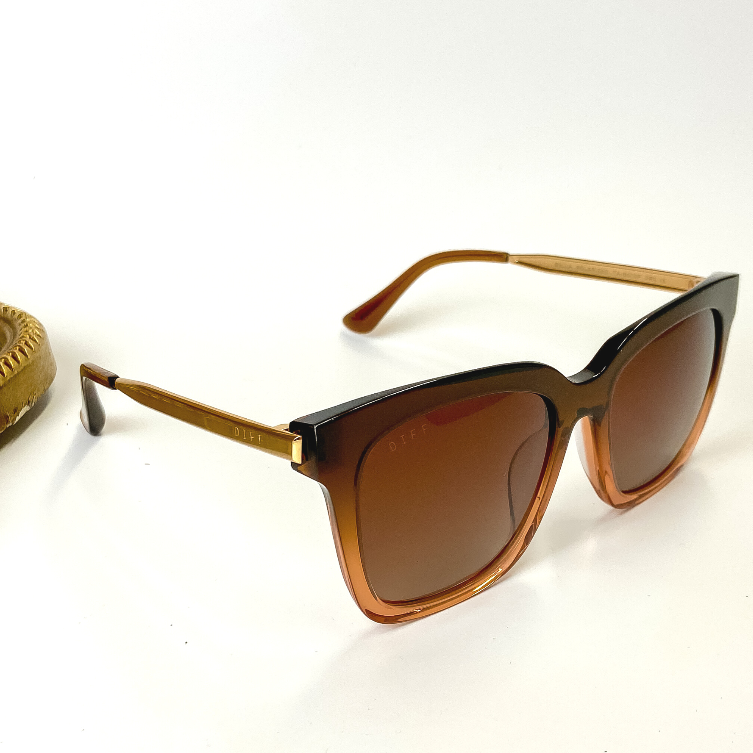 A pair of brown lens sunglasses with taupe ombre frames. These sunglasses are pictured on a white background with gold jewelry.