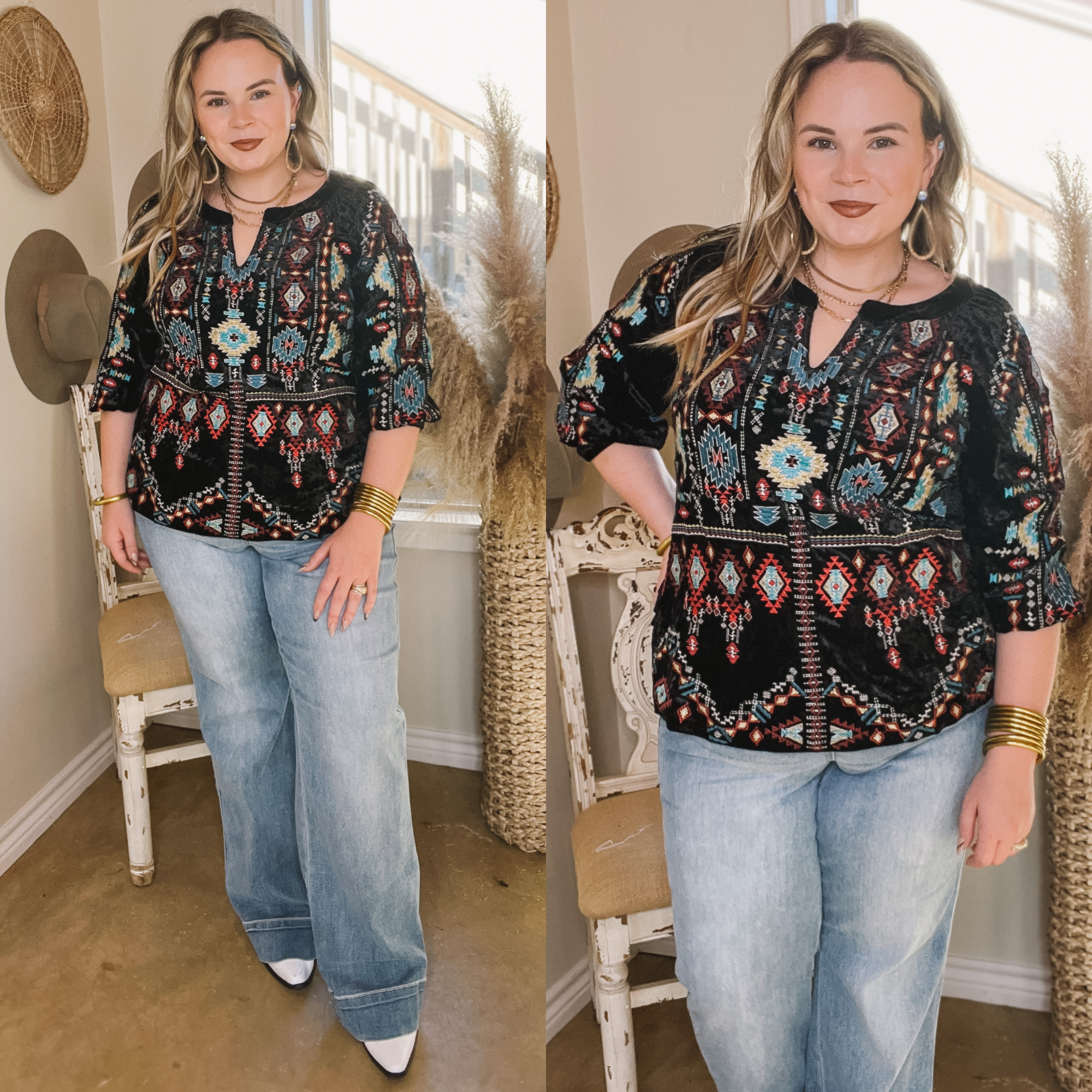Model is wearing a black embroidered velvet top with 3/4 length sleeves. The embroidery on this top includes the colors red, blue, maroon, yellow and white. Model has this top paired with light wash trouser jeans, white booties, and gold jewelry. Background is hues of white and brown. 