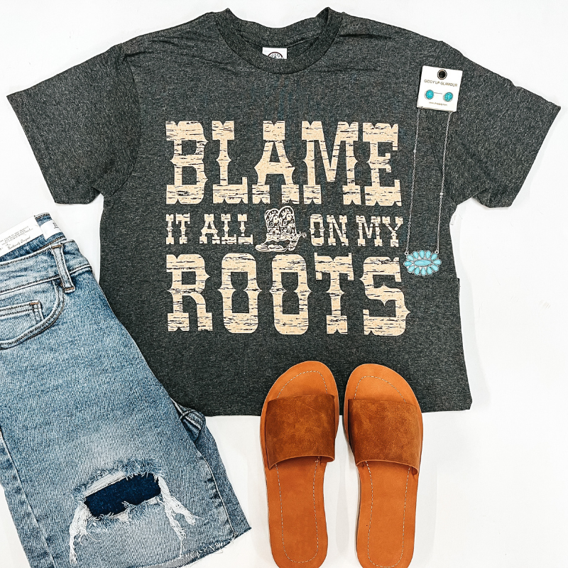 A grey tee shirt that says "Blame It All On My Roots I Showed Up in Boots". Pictured with tan sandals, denim shorts, and turquoise jewelry.