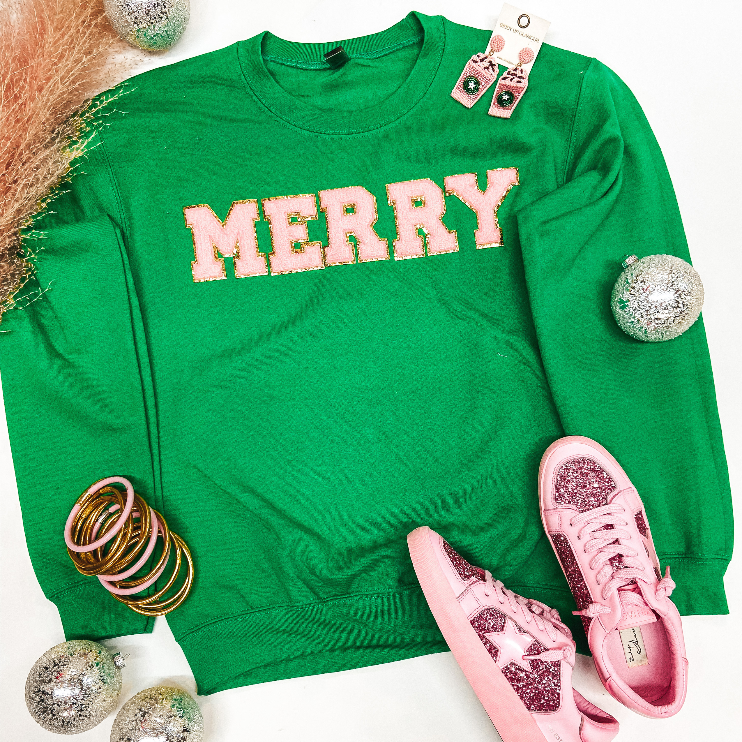 A long sleeve crew neck sweatshirt that is bright green with "Merry" across the chest in pink chenille letters with gold trim. Pictured on a white background with sneakers, gold and pink jewelry, and Christmas ornaments. 
