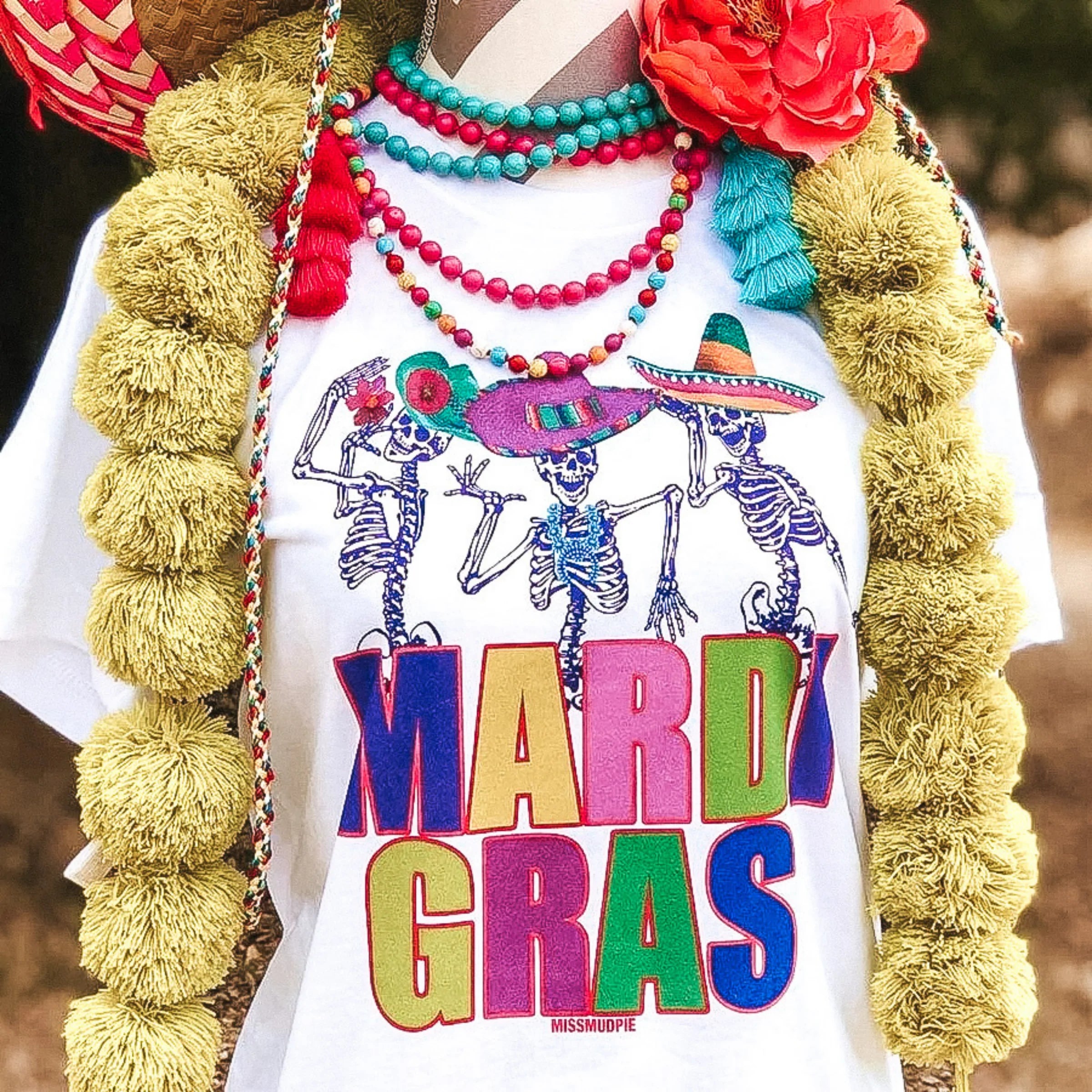 A white crew neck tee shirt with short sleeves. This tee has a graphic of dancing skeletons that says "Mardi Gras" in lots of colors.