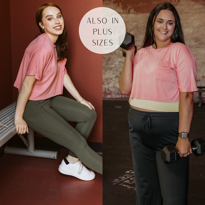 Models are wearing a coral short sleeve crop top with a front pocket. Size small model has it paired with olive green leggings and white sneakers. Plus size model has it paired with a yellow sports bra and black sweatpants.