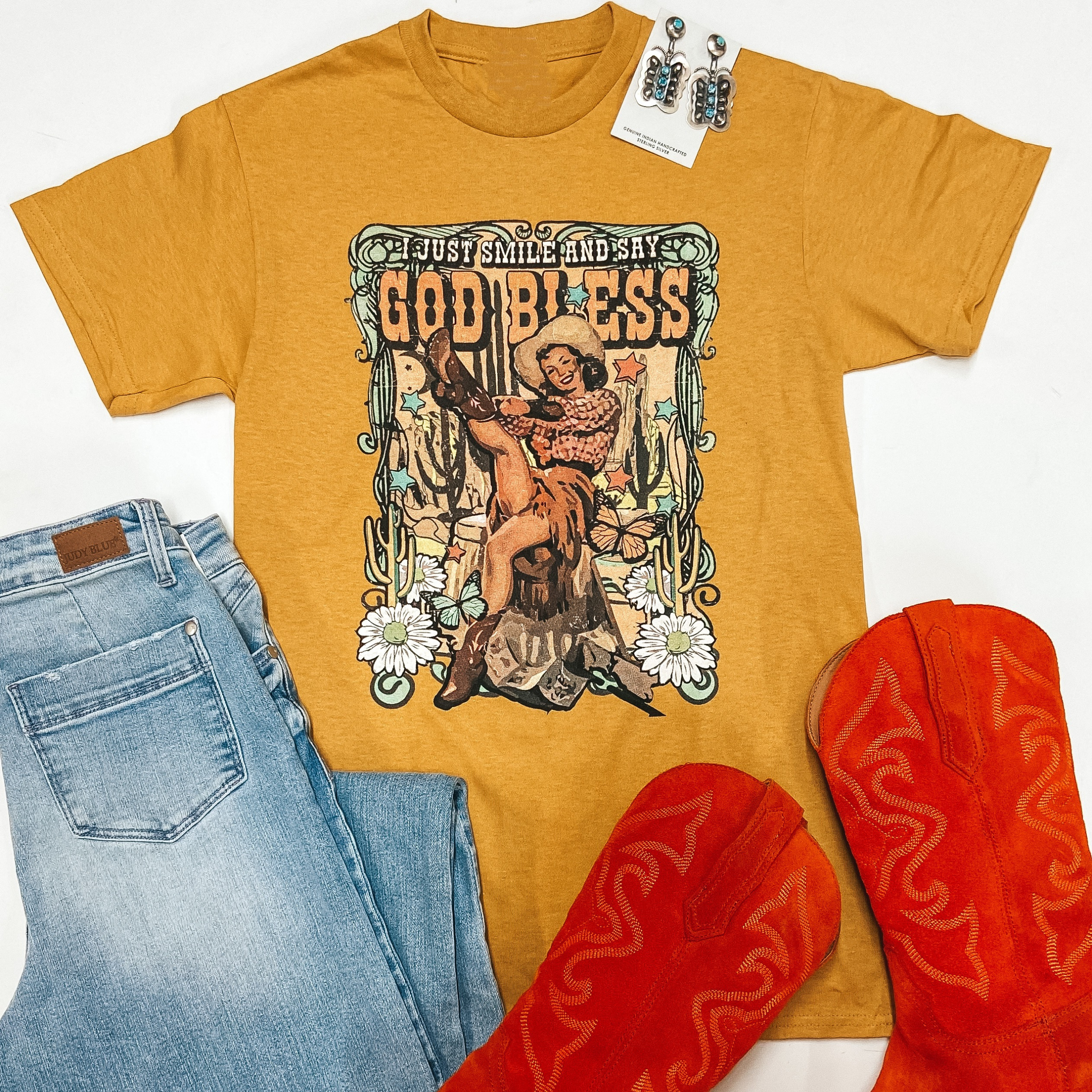 A mustard yellow short sleeve tee shirt is pictured on a white background. On the center of the tee is a graphic of a pin up cowgirl that says "I just smile and say God Bless." It is pictured with orange boots. light wash jeans. and sterling silver earrings.