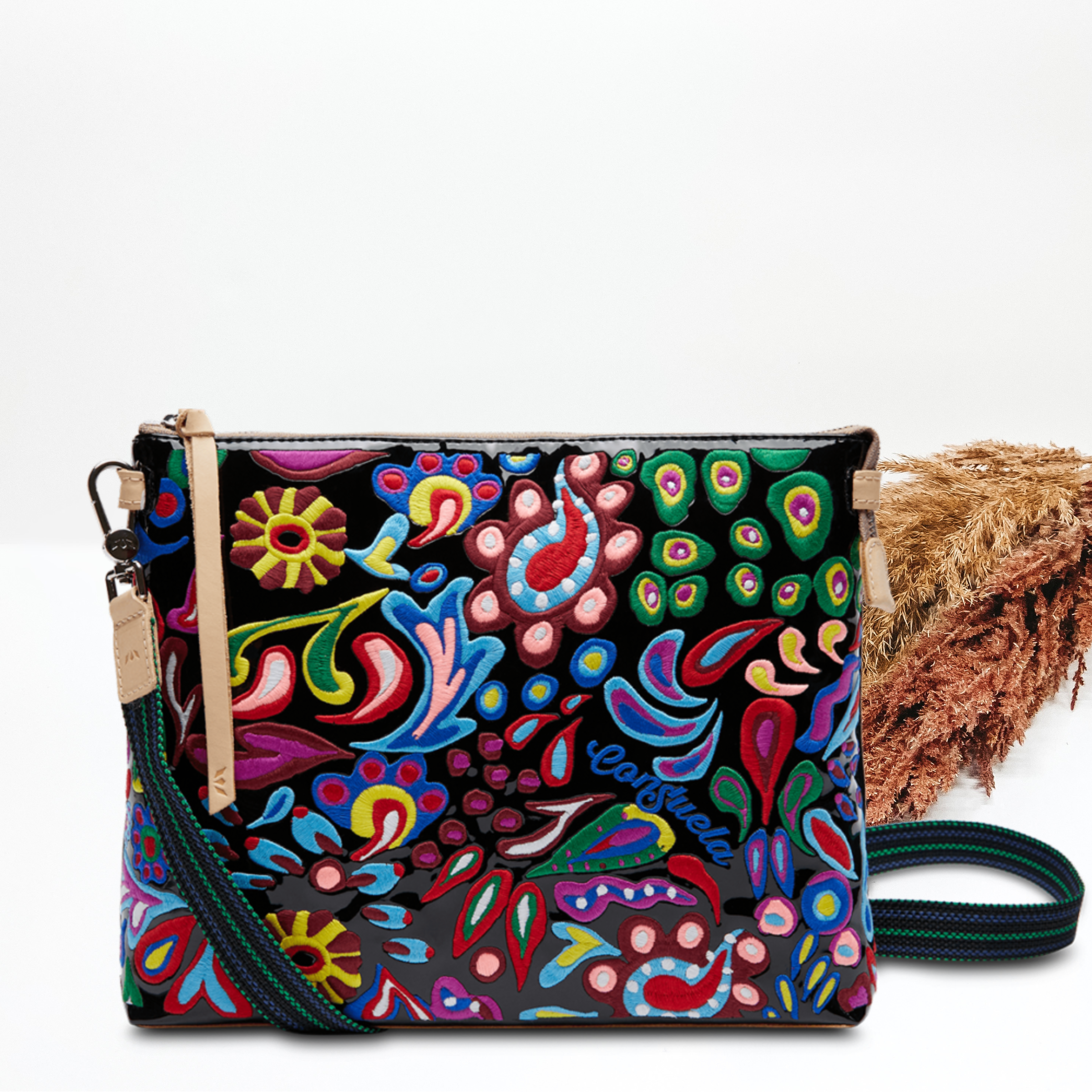 Black rectangle purse with a multicolored embroidered design. This bag also has tan leather detailing and a striped purse strap. This purse is pictured on a white background with tan and brown pompous grass in the background.