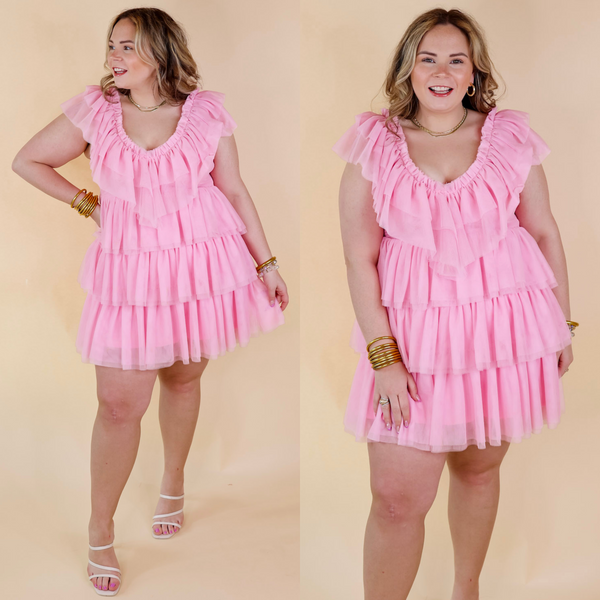 Model is wearing a light pink short dress made of ruffle tulle. Model has this dress paired with white strappy heels and gold jewelry.