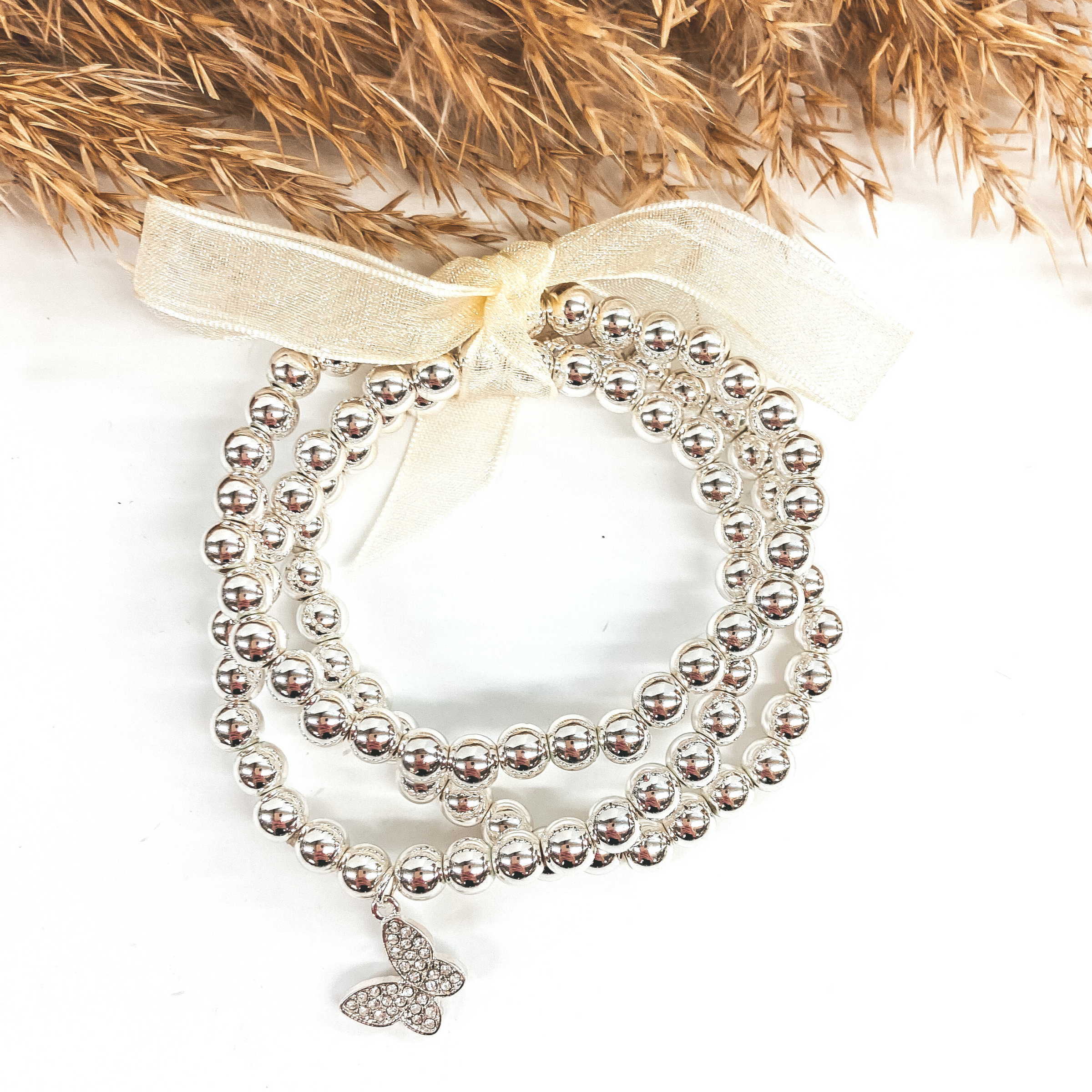 Three silver circle bracelets with a crystal butterfly charm. Pictured on a white background with pampus grass.