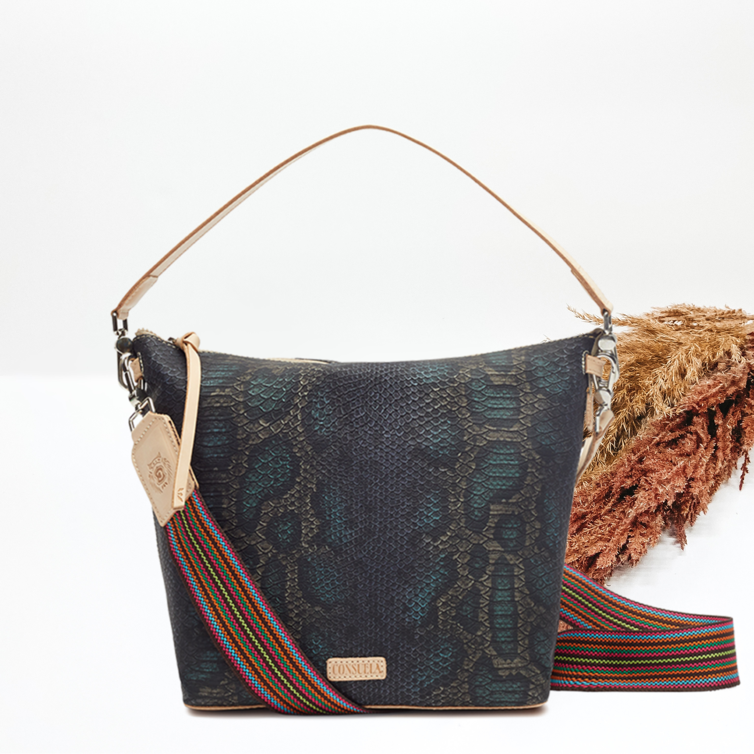 This bag has a smaller base with a wider top. This a dark blue and black snake print bag. This bag also has a striped handle and a wide tan leather strap. This bag is pictured on a white background with tan and brown pompous grass in the background.