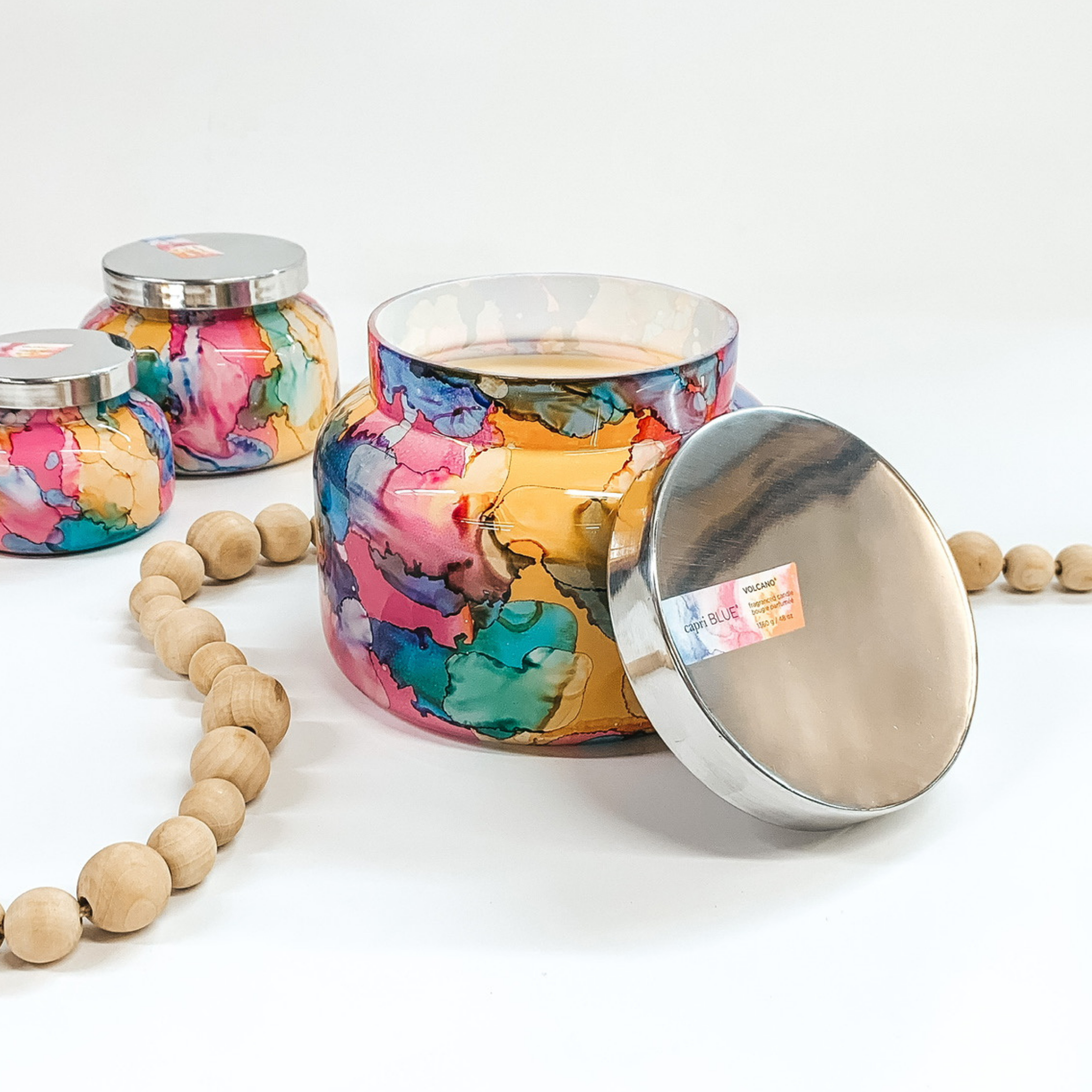 A rainbow watercolor glass jar candle with a metal lid. Pictured on white background with wooden beads and smaller candles.