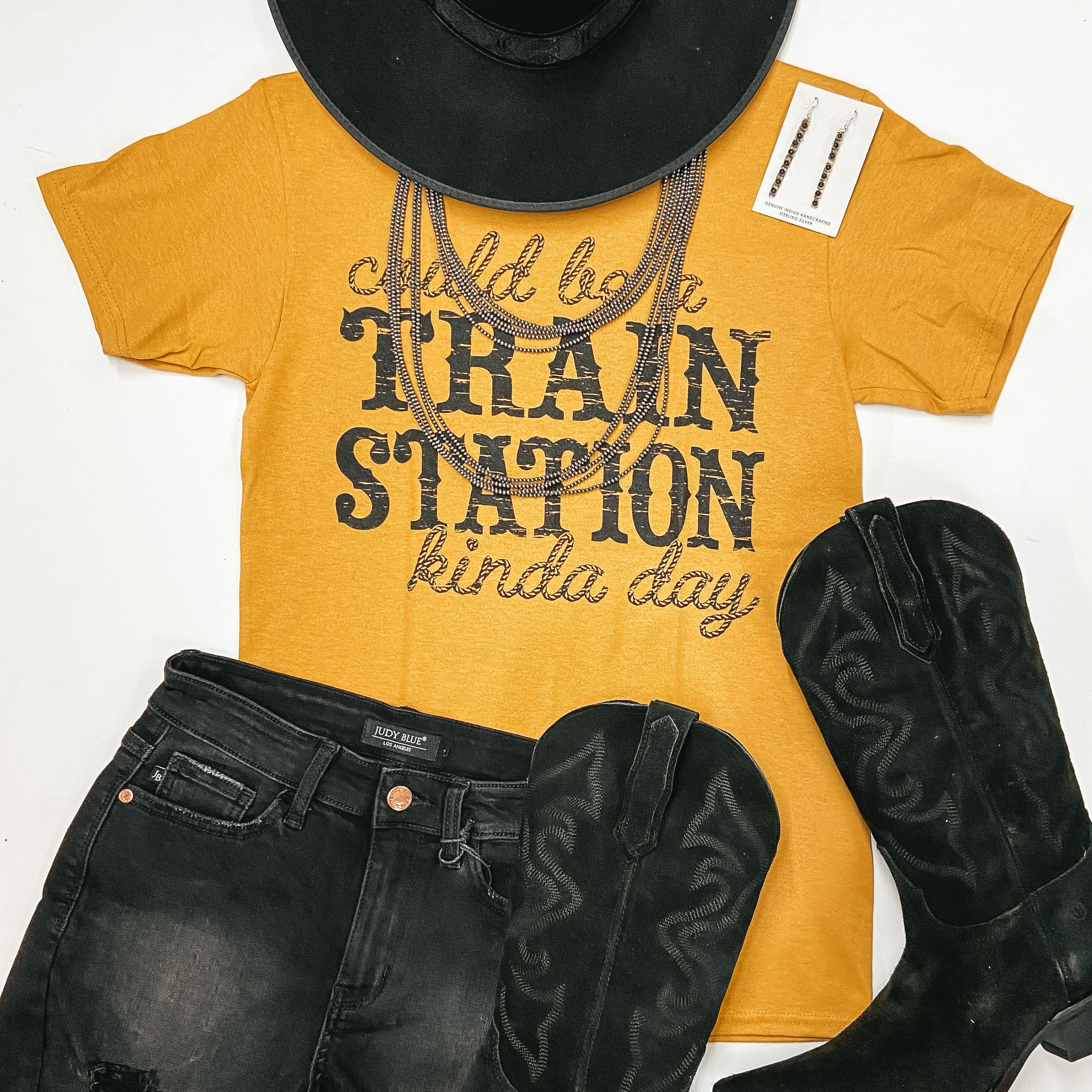 A mustard yellow graphic tee that says "Could Be A Train Station Kinda Day." Pictured on a white background with a black hat, black shorts, and silver jewelry.