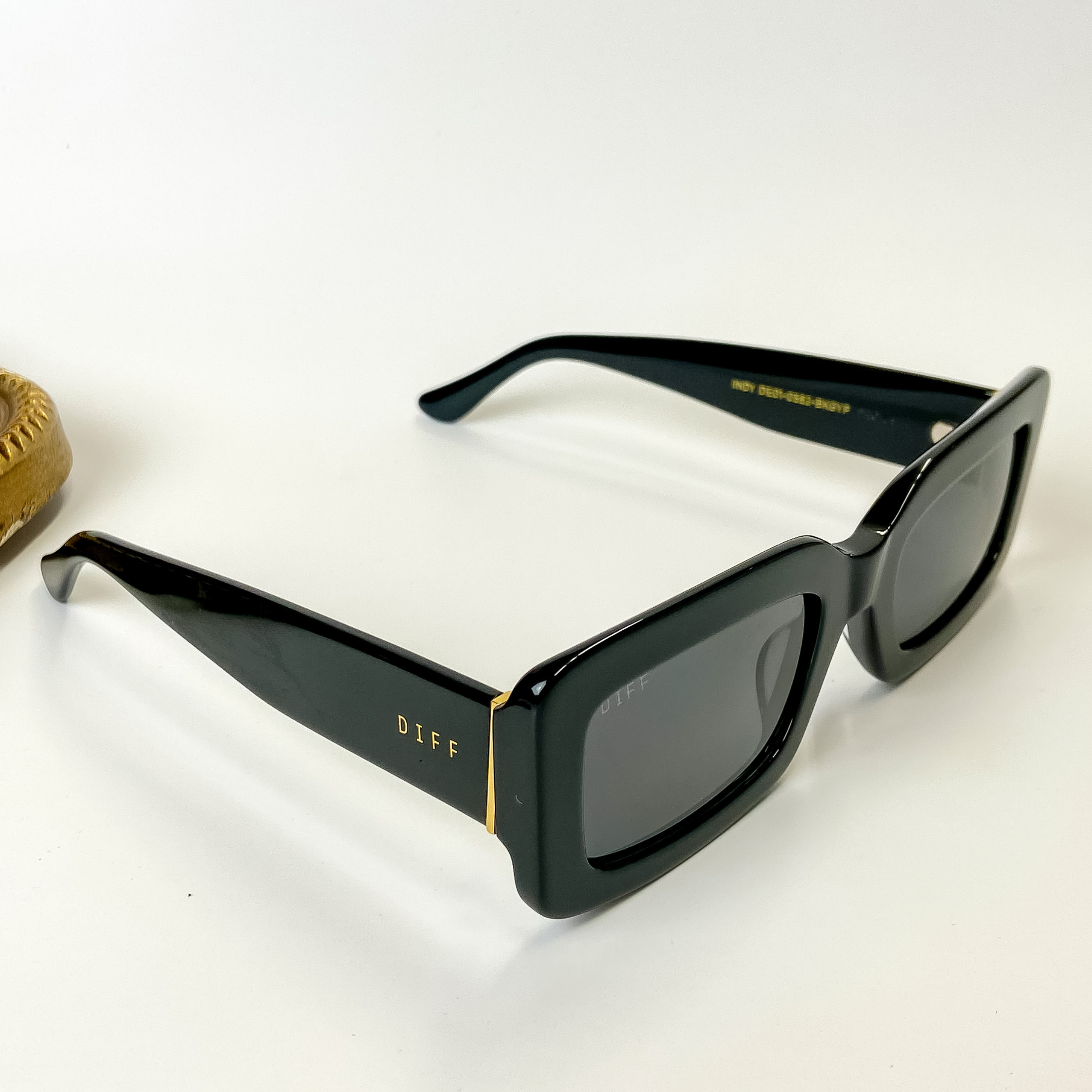 A pair of black sunglasses with rectangle lenses. Pictured on a white background with gold jewelry.