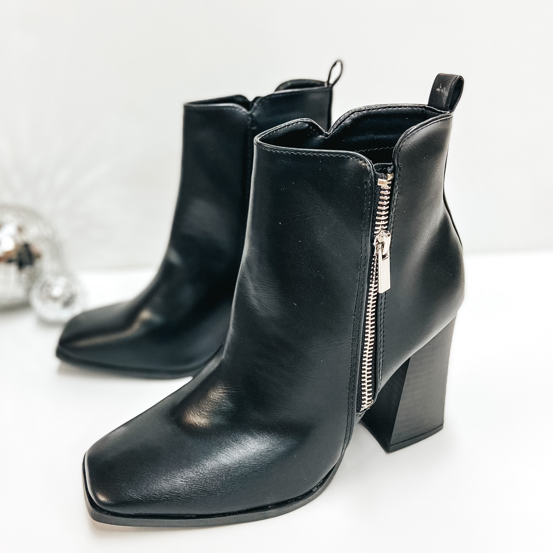A pair of black zip up booties that have a high heel and square toe. Pictured on white background with disco balls.