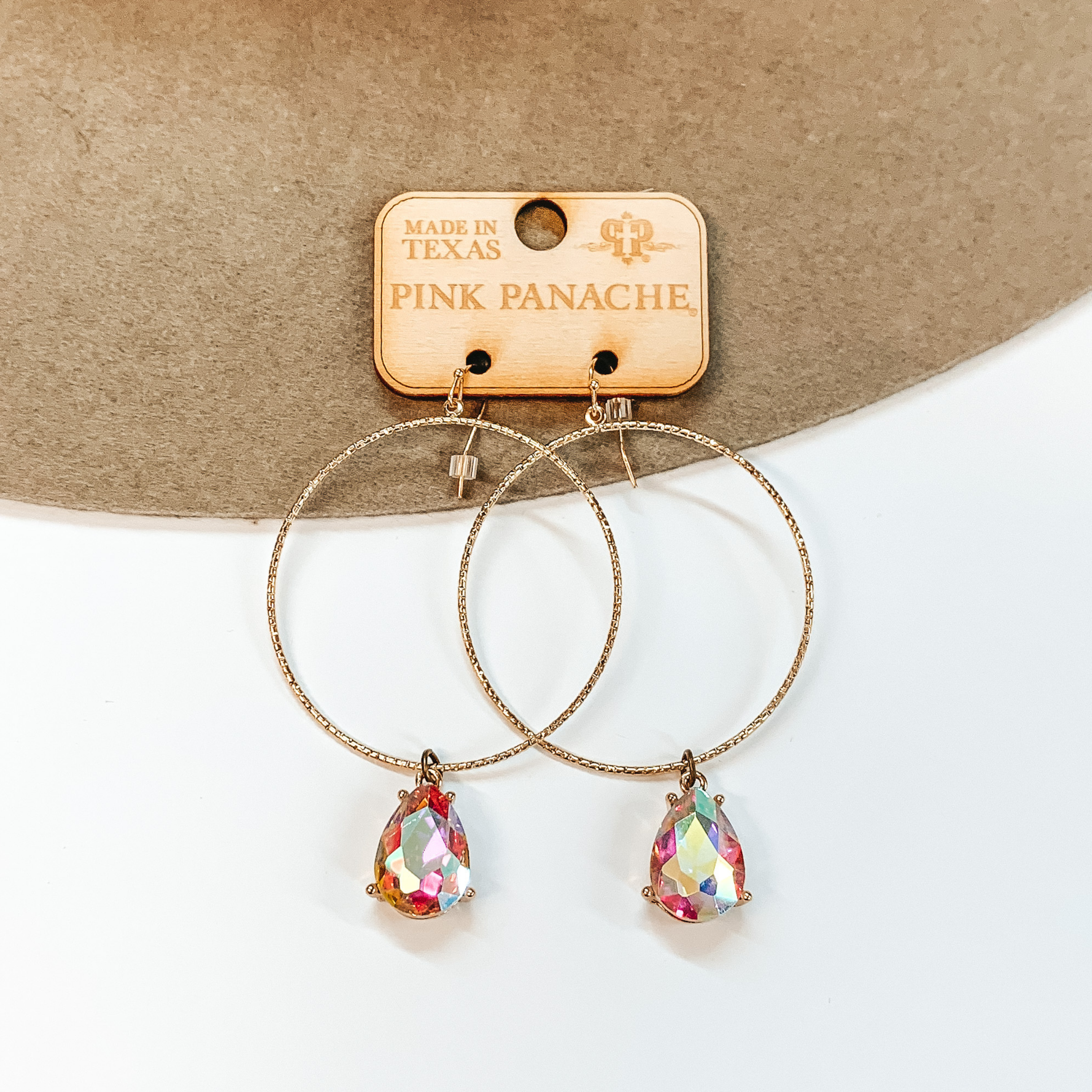 A pair of textured gold hoop earrings that have a colorful crystal dangle. Pictured on a beige hat on a white background.