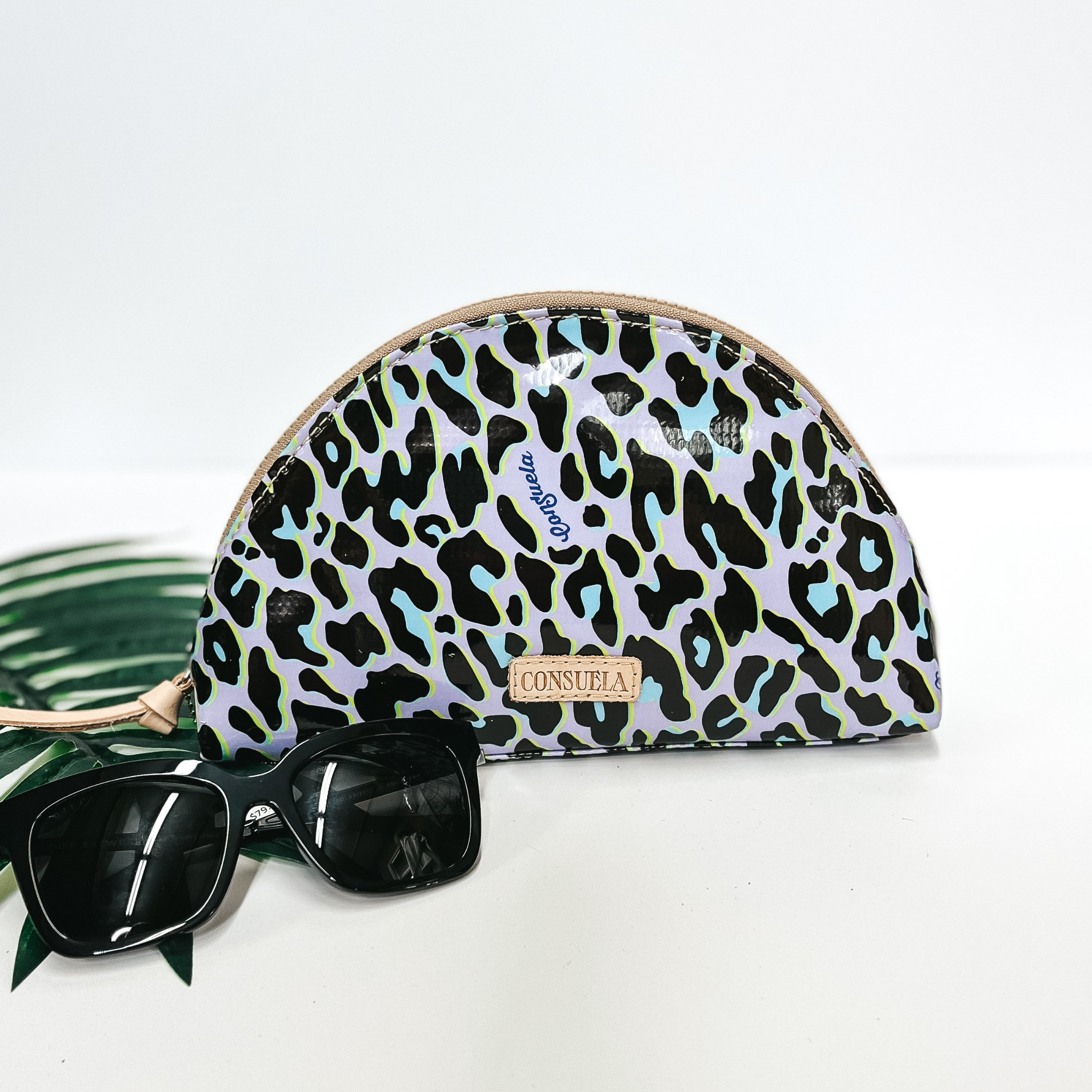 A purple leopard print dome cosmetic bag with leather zipper. Pictured on white background with a palm leaf and black sunglasses.
