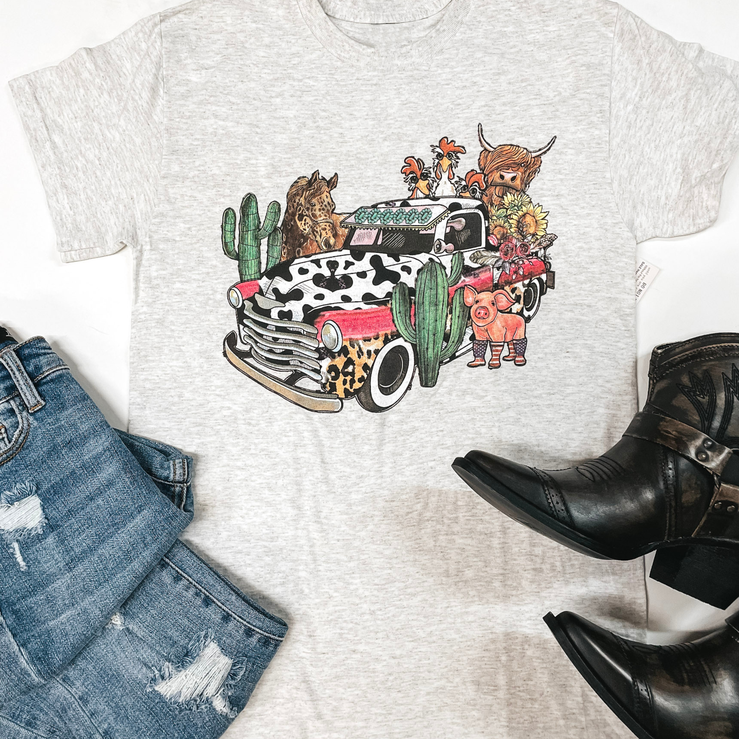 A gray tee is in the center of the picture on a white background. In the center of the tee is a cow-print truck loaded with cows, chickens, pigs and sunflowers. Cactus stands to the left of the truck. Light wash jeans are placed on the bottom left side of the picture and black boots are placed on thee bottom right of the picture/. 