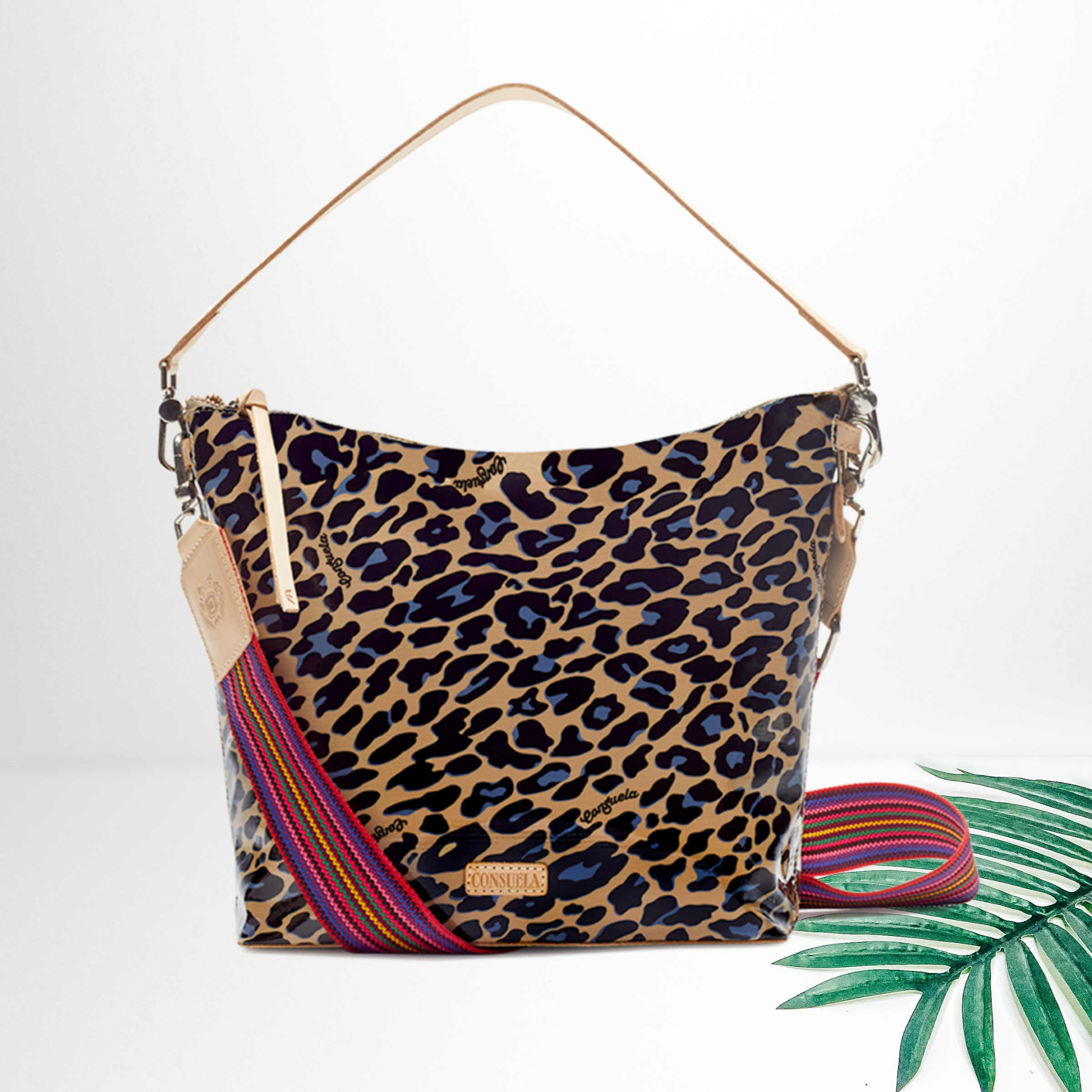 Centered in the picture is a medium sized bag in a leopard print with hints of blue. To the right of the bag is a palm leaf, all on a white background. 