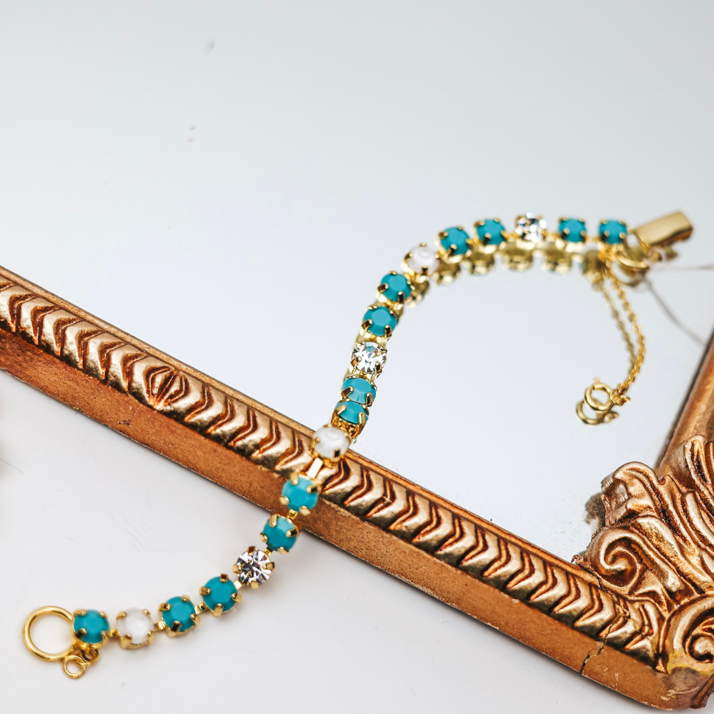 A gold-tone bracelet with a mix of ivory, clear, and turquoise crystals along the bracelet. Pictured on a white background with a gold mirror.