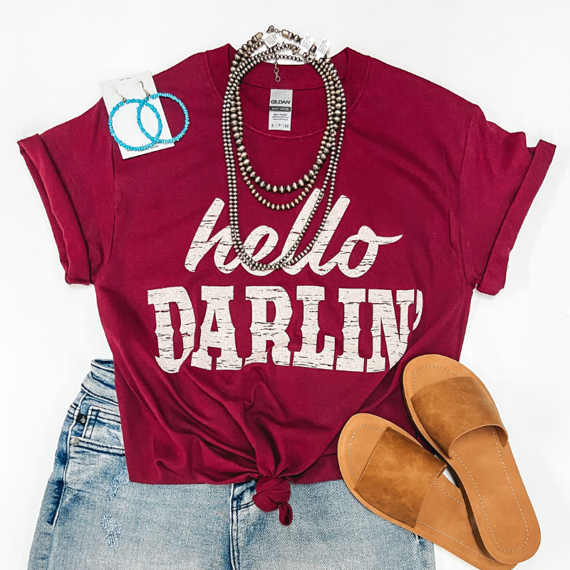 A maroon tee shirt that says "Hello Darlin'" in ivory letters. Pictured with genuine turquoise jewelry, tan sandals, and denim shorts.
