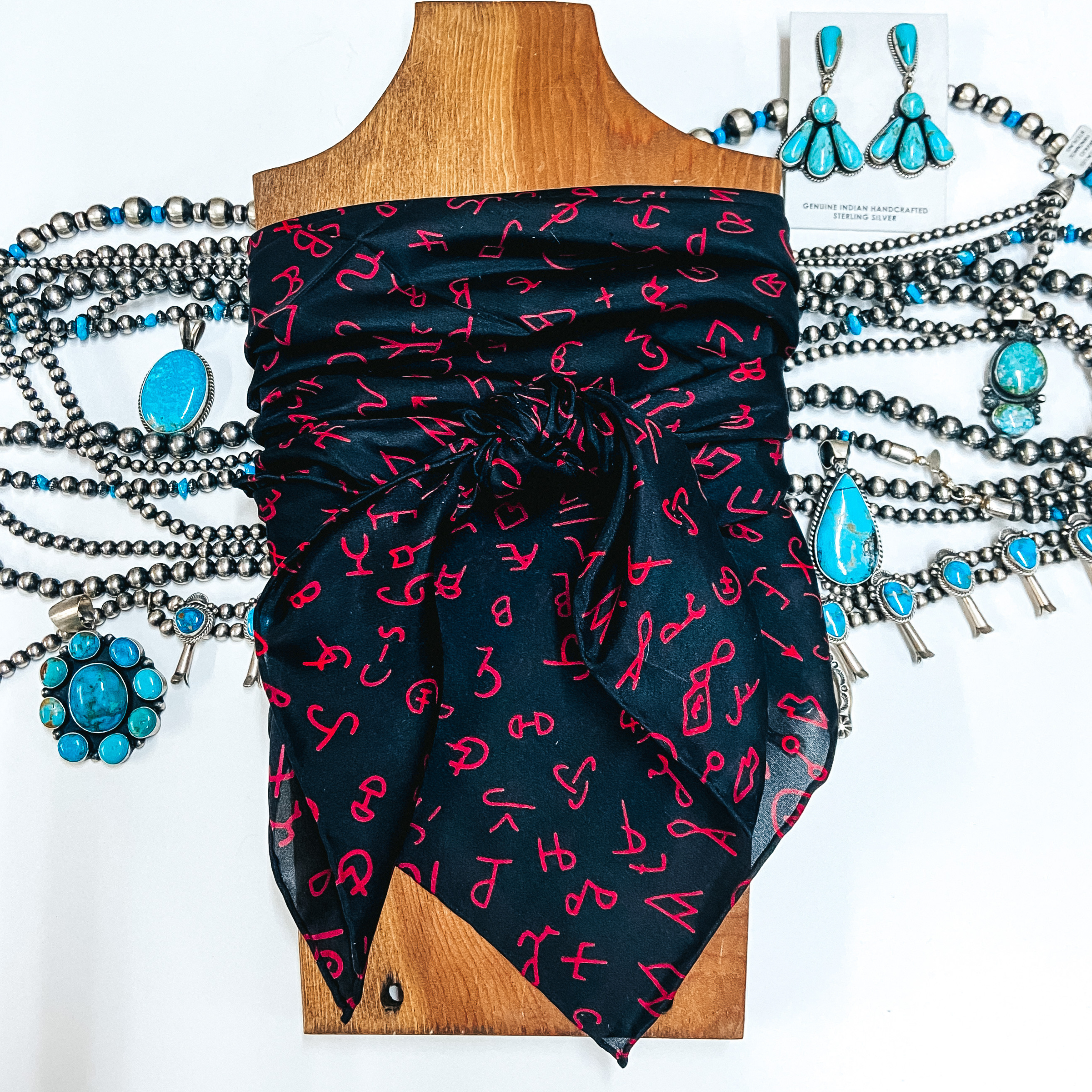 A silky scarf with branded patterns tied on a wooden display. Pictured on white background with turquoise and silver jewelry.