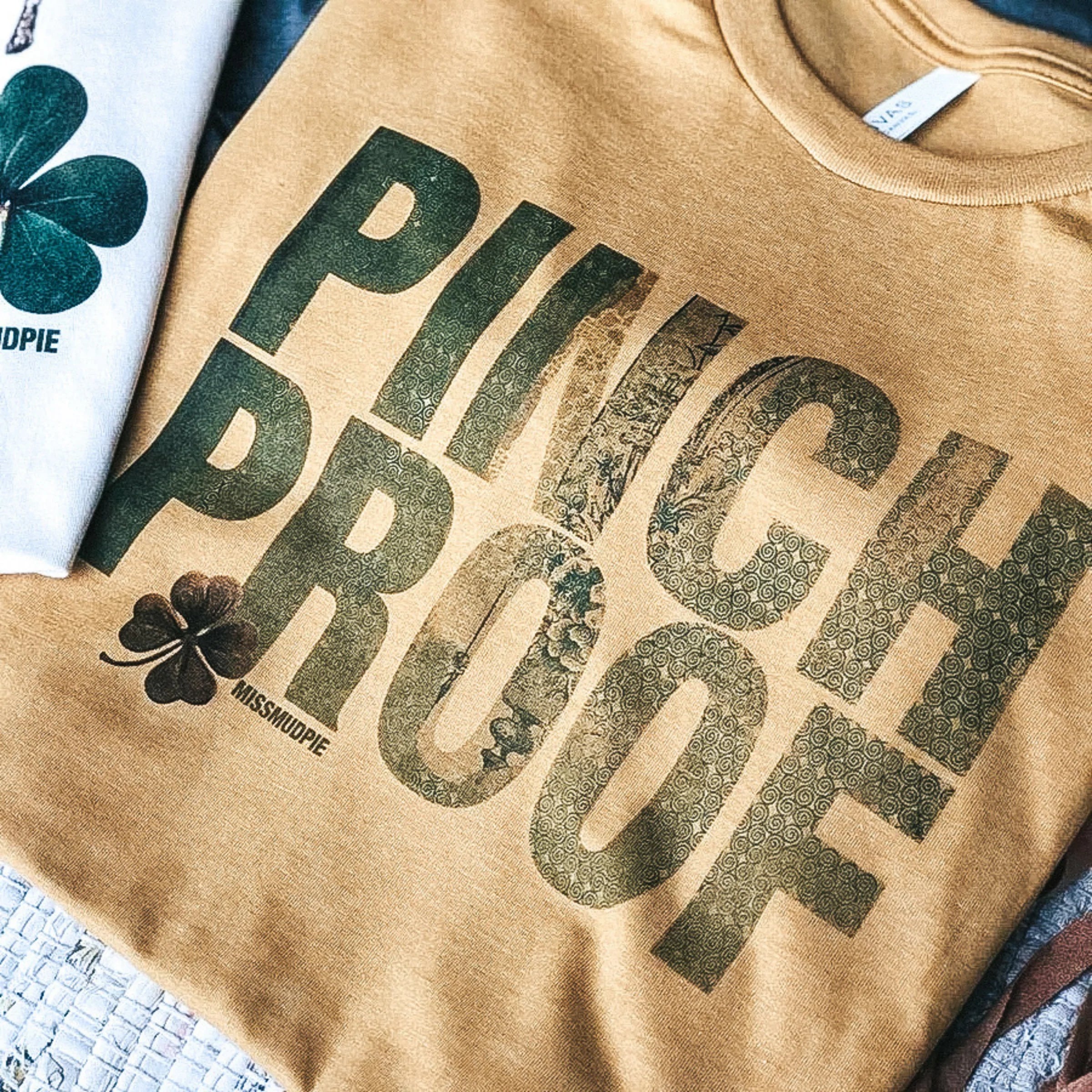 An army green colored tee shirt folded exposing the graphic that says "Pinch Proof" in olive green.