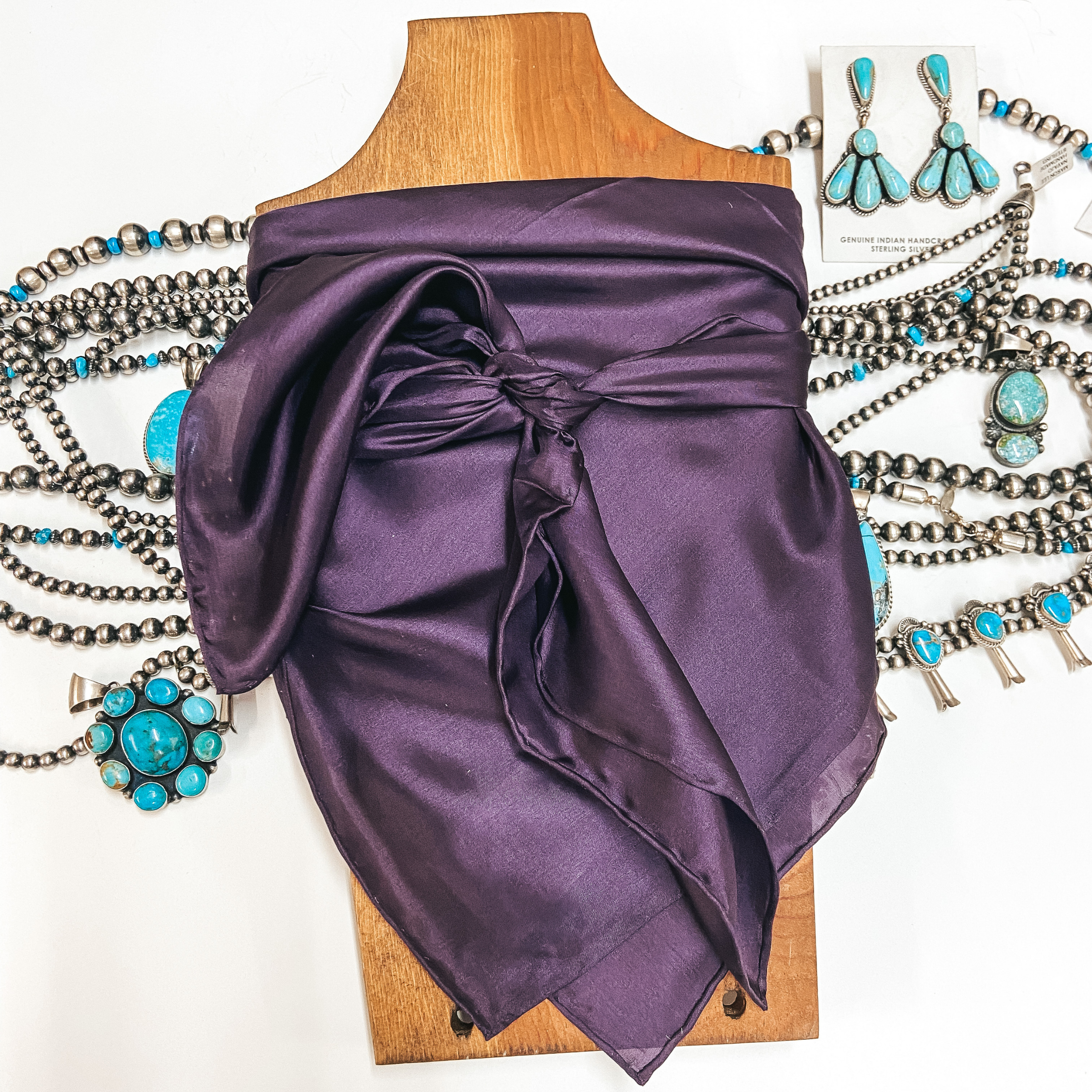 A solid color wild rag tied around a wooden display. Pictured on white background with turquoise jewelry.
