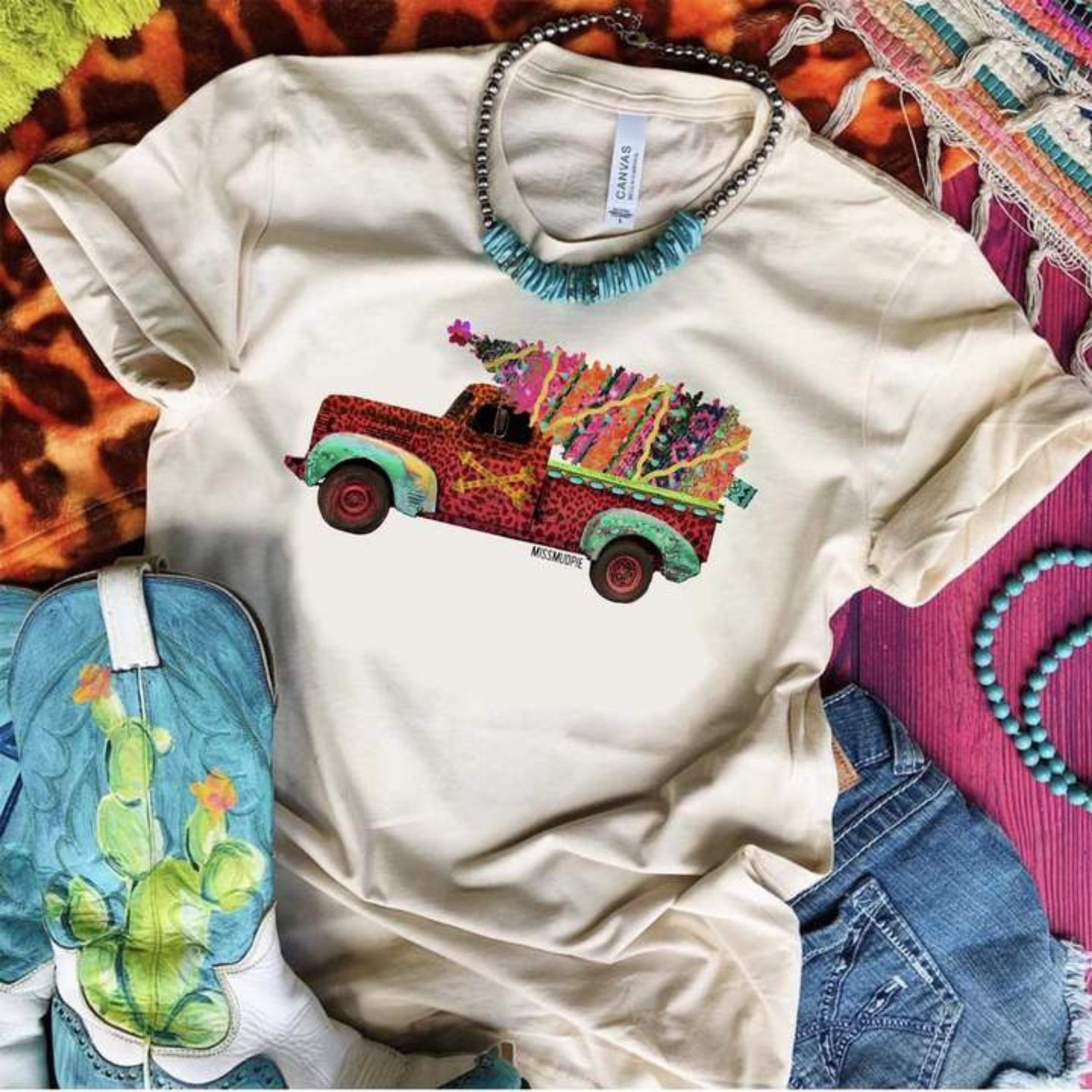 This Bella + Canvas cream tee includes a crew neckline, short sleeves, and a cute Christmas graphic of a red vintage truck and colorful Christmas tree in the back. This tee is shown as a flat lay with rolled sleeves, turquoise necklace, and turquoise cowboy boots. 