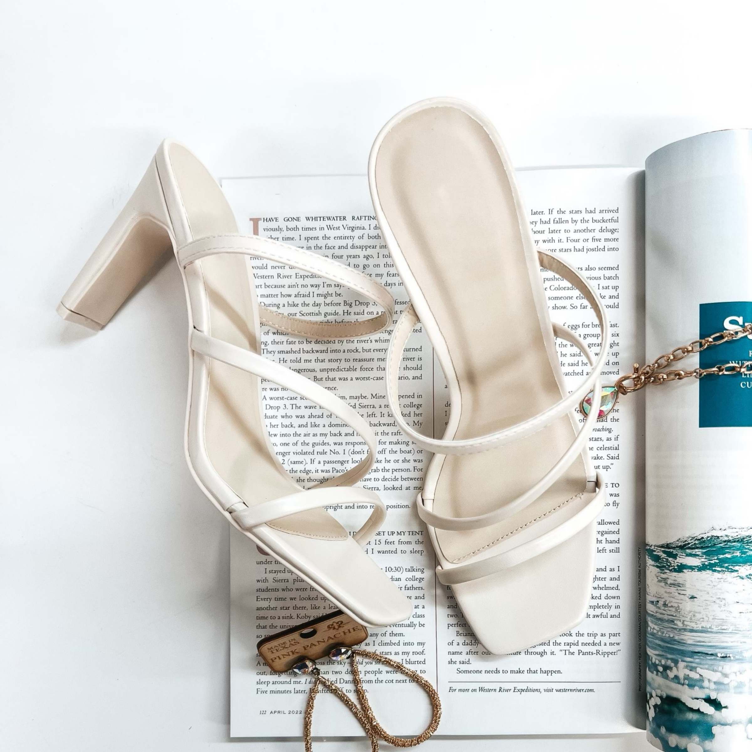 A pair of ivory strappy heels that have a square toe. These shoes are pictured on a white background with gold jewelry and a magazine.