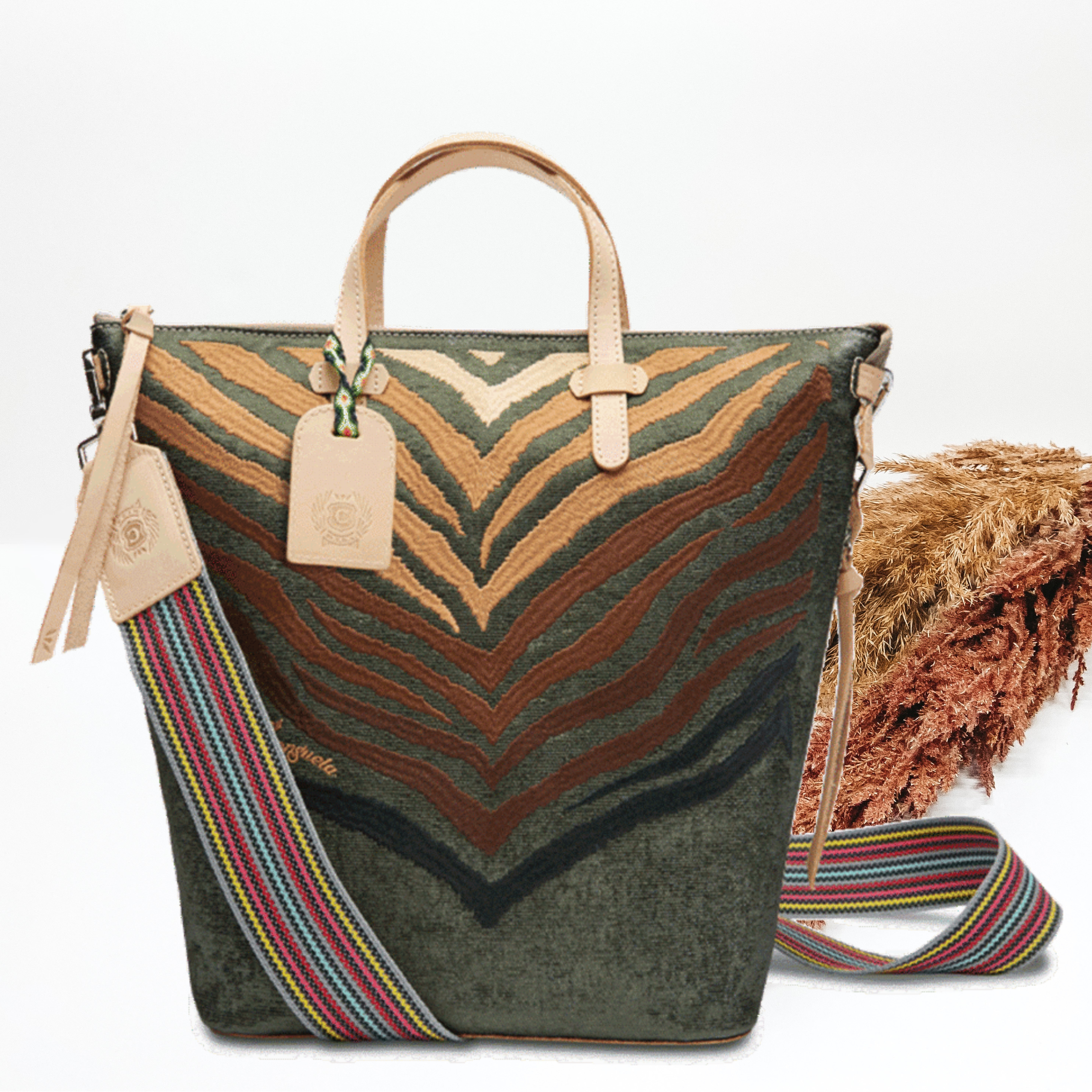 This bag has a smaller base with a wider top. This is a dark green bag that has an embroidered zebra print. The zebra print goes from an ivory color to a black color. This bag has small, leather tan handles at the top and a striped strap on the sides. This bag is pictured on a white background with tan and brown pompous grass in the background. 