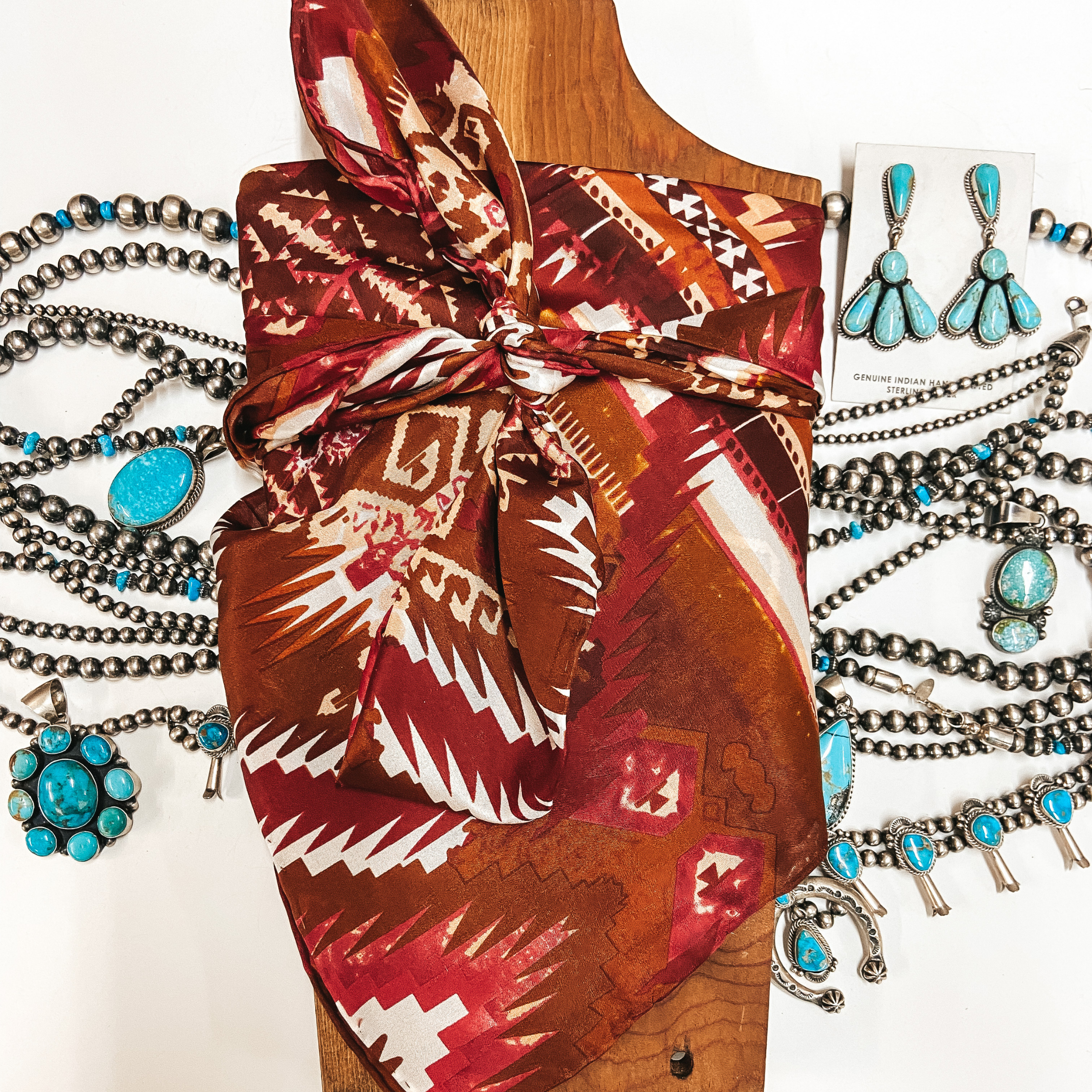 An Aztec print scarf with red, brown, and white colors. Scarf is tied around a wooden display with turquoise and silver jewelry.