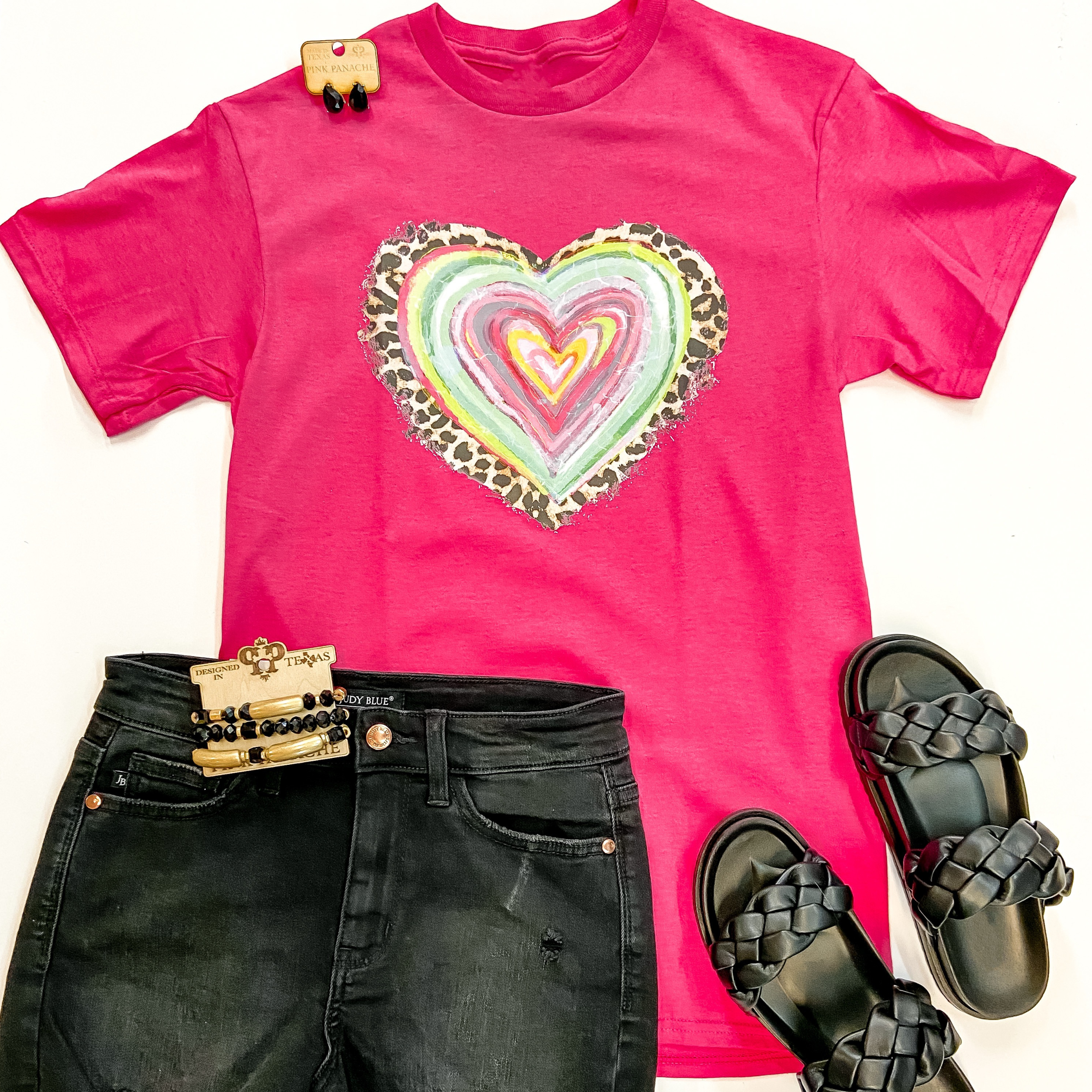 A hot pink tee shirt with short sleeves, a crew neckline, and a heart graphic at the center that is leopard print, pink, green, and yellow. This tee is pictured on a white background with black shorts, black sandals, and black Pink Panache jewelry.