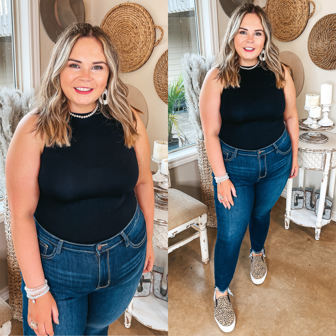 Model is wearing a high neck tank top bodysuit that is black. Model has it paired with dark wash skinny jeans, leopard print sneakers, and ivory jewelry.