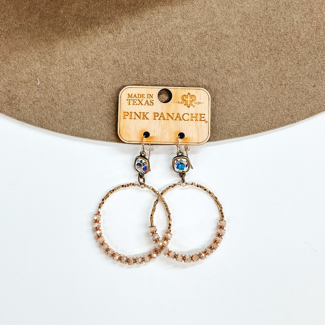 AB cushion crystals with a hanging open circle pendant. The circle pendant includes gold and ivory beads. These earrings are pictured on a brown and white background. 