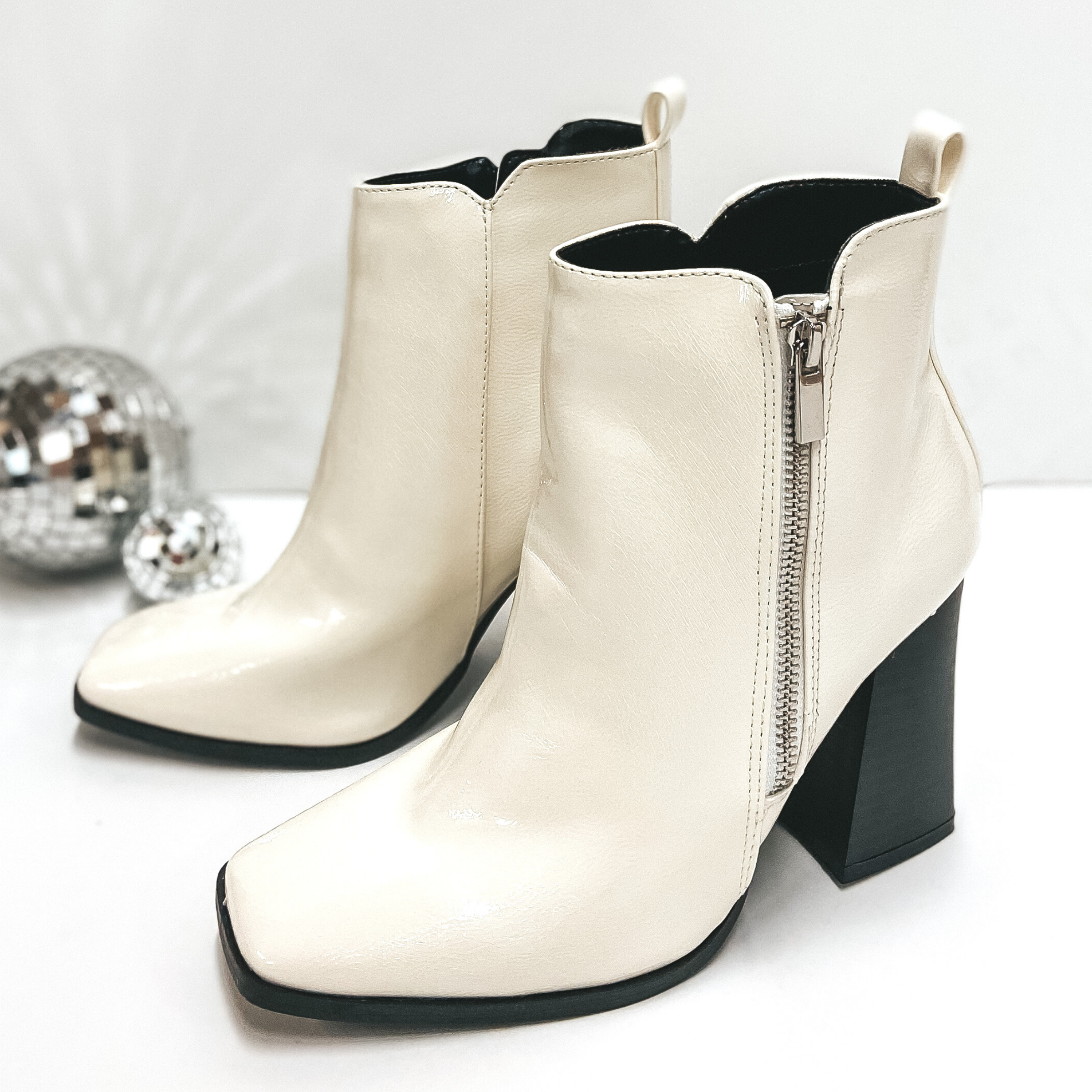 A pair of ivory booties with a square toe and a black high heel. Pictured on white background with disco balls.