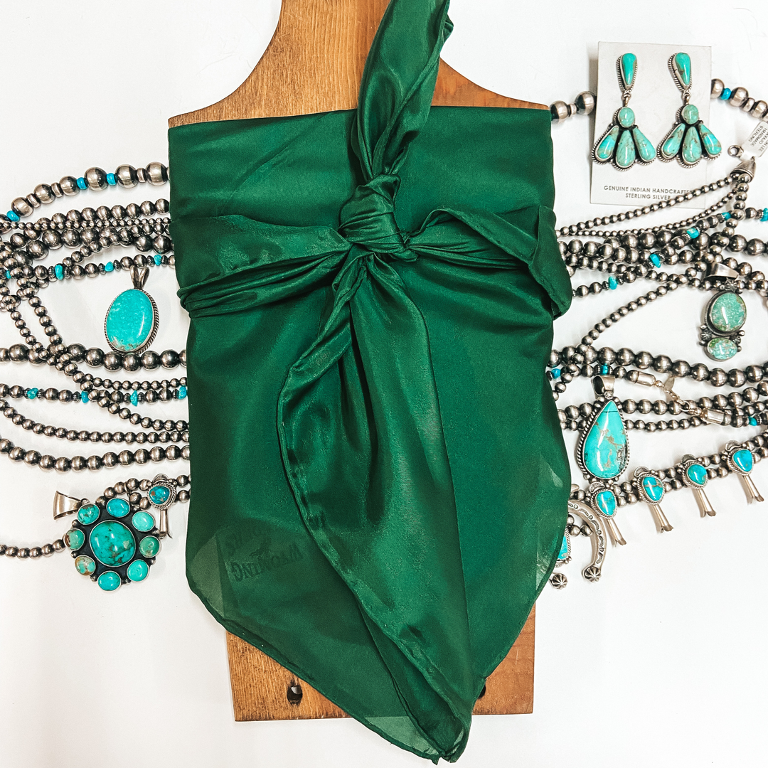 A solid color wild rag tied around a wooden display. Pictured on white background with turquoise jewelry.