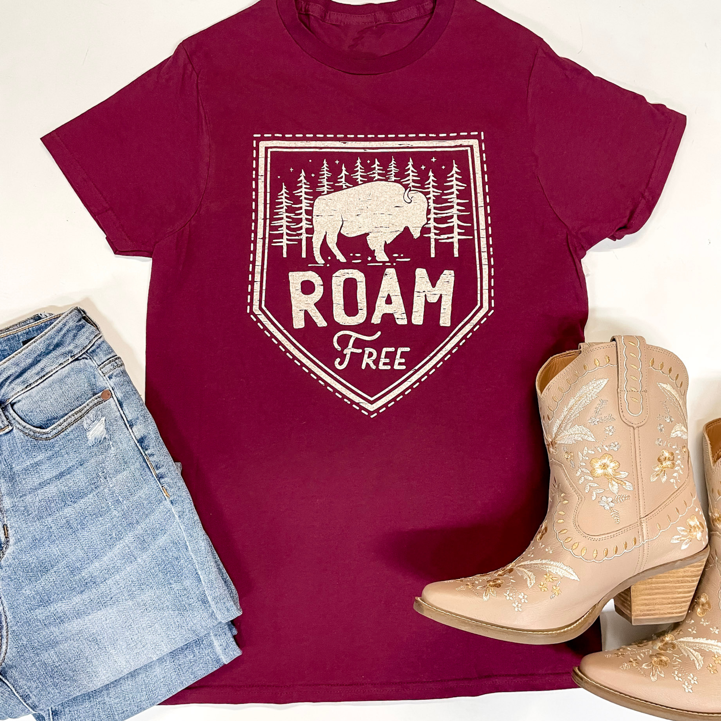 A maroon tee is centered in the middle of the picture on a white background. In the middle of the tee is a buffalo on top of block letters that state, "roam free." On the bottom left of the picture is light-wash jeans and on the bottom right is cream colored boots. 