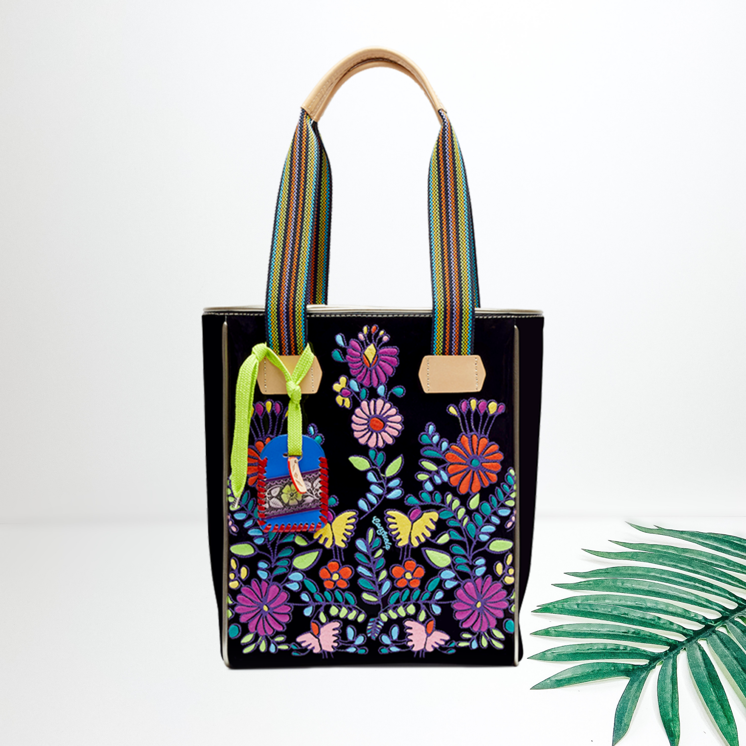 Centered in the picture is a tote bag in black with multi colored embroidery. To the right of the tote is a palm left, all on a white background.