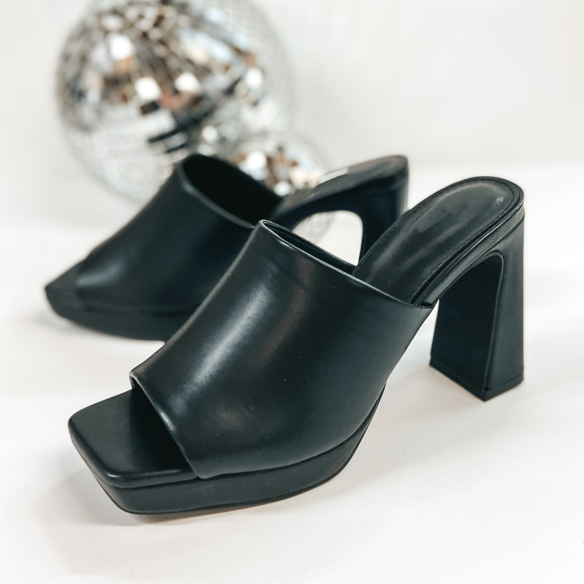 A pair of platform shoes with a slip on upper and open toe. These shoes have a high block heel. The black heels are pictured on white background with disco balls.