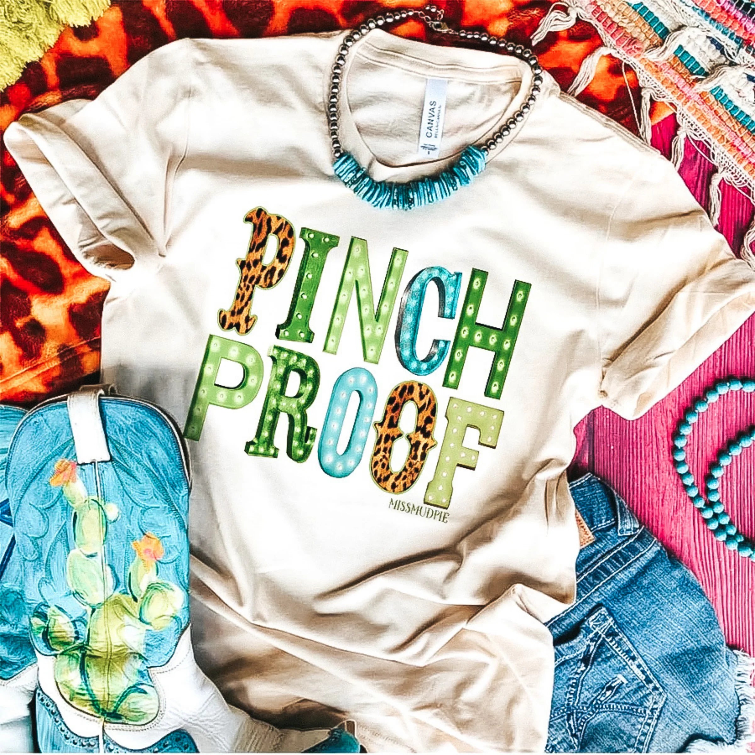A cream colored tee shirt with a multi print graphic that says "Pinch Proof." Pictured on pink background with cowgirl boots and turquoise jewelry.