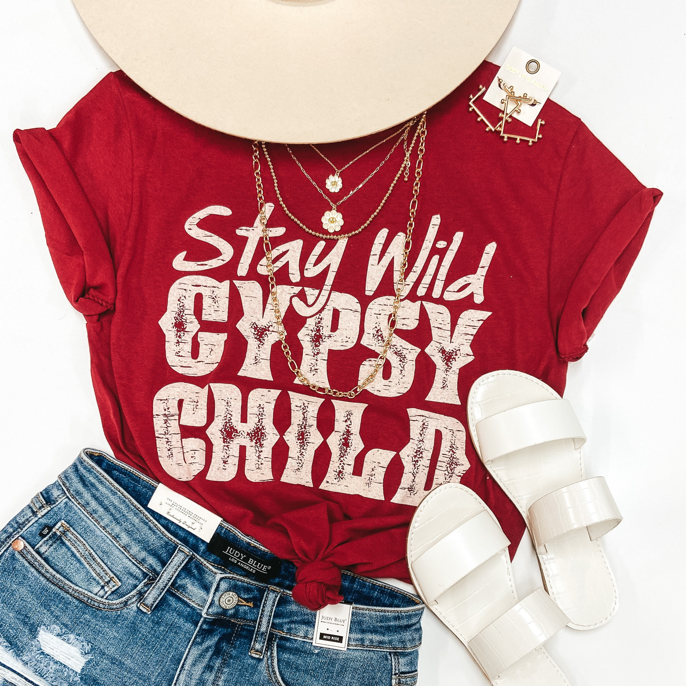 A red graphic tee with cuffed sleeves and a knotted front. The tee shirt has a graphic that says "Stay Wild Gypsy Child" in ivory writing. Tee shirt is pictured on white background with denim shorts, gold jewelry, white sandals, and an ivory hat.