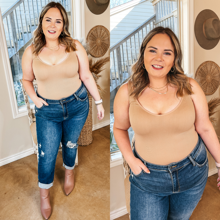 Model is wearing a nude tank top bodysuit with a deep v plunge. Model has it paired with distressed boyfriend jeans, taupe booties, and gold jewelry.