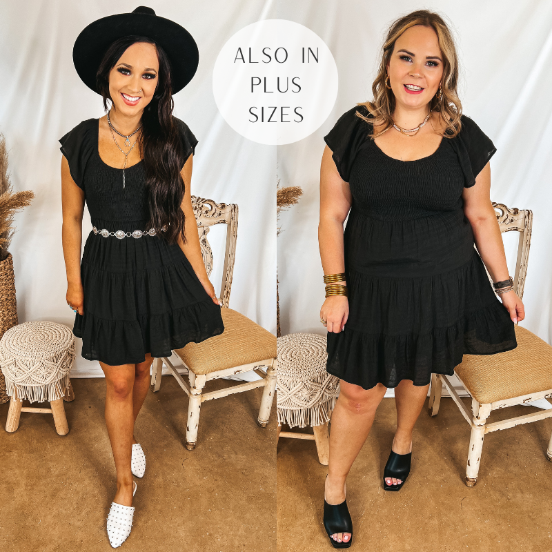 Models are wearing a black dress that has ruffle cap sleeves, a smocked bodice, and a tiered skirt. Size small model has it paired with white mules, a silver belt, and a black hat. Size large model has it paired with black heels and gold jewelry.