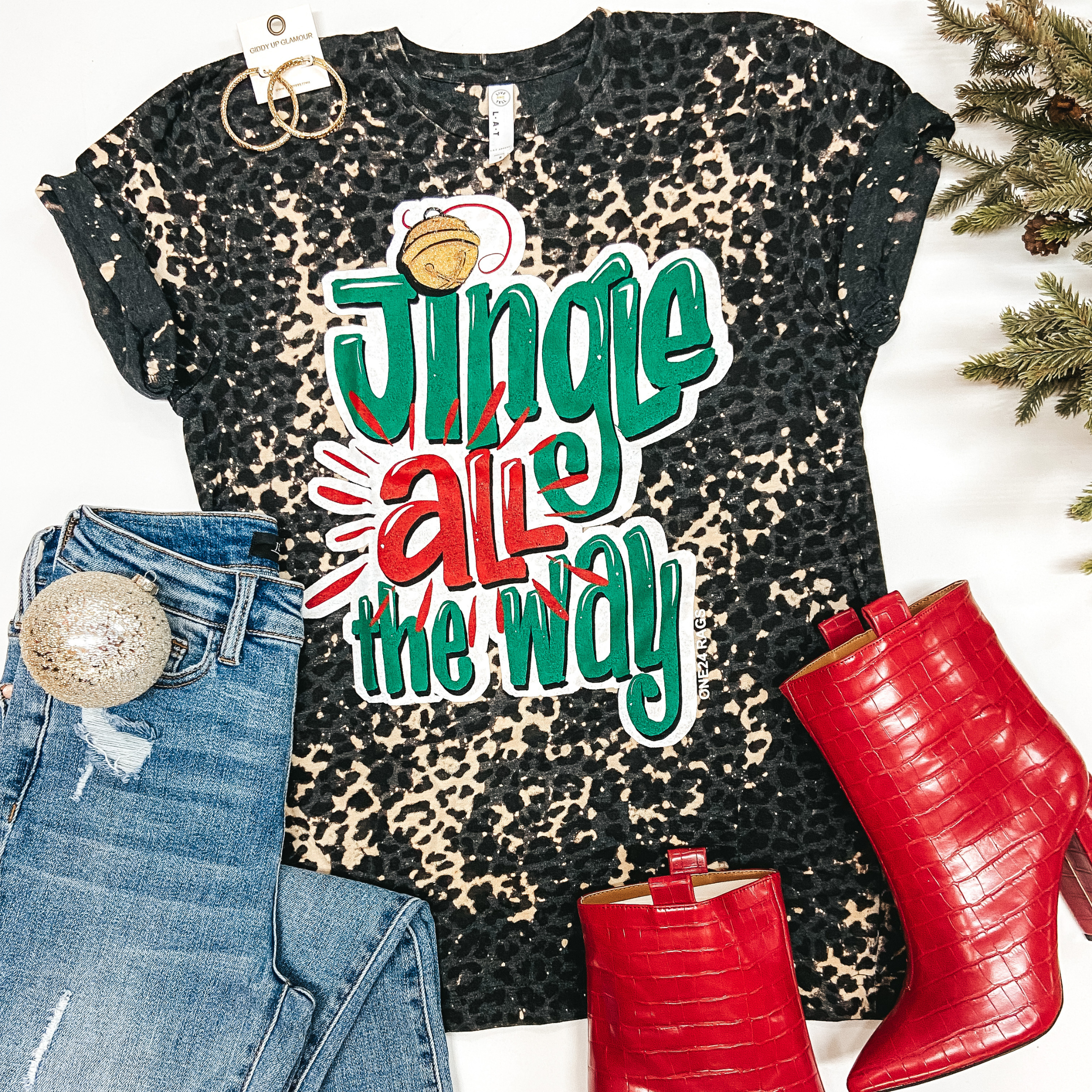 A charcoal grey tee shirt with short sleeves and a crew neckline. This tee shirt has bleach splatter detailing underneath a black faux leopard print. The graphic on the front says "Jingle All The Way." Pictured on a white background with gold jewelry, red booties, and Christmas decor.