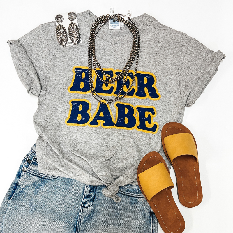 A grey tee shirt that says "Beer Babe" on the front in yellow and navy letters. Pictured on white background with yellow sandals, denim shorts, and sterling silver jewelry.
