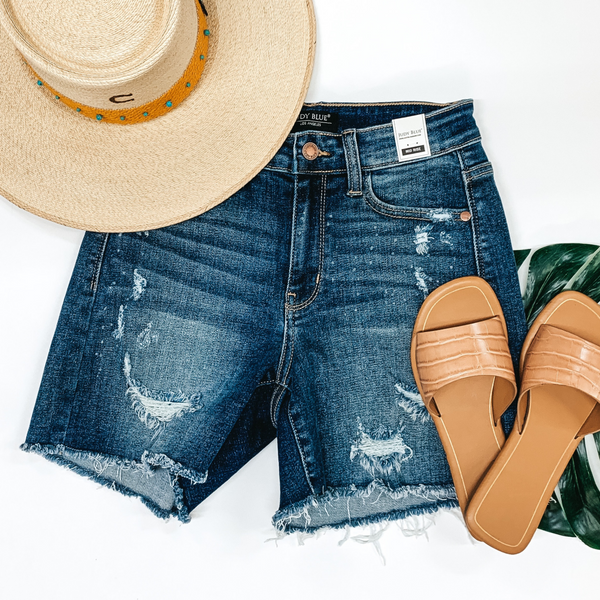 A pair of ripped and bleach distressed shorts with a frayed hem. Pictured on white background with a straw hat and tan sandals.